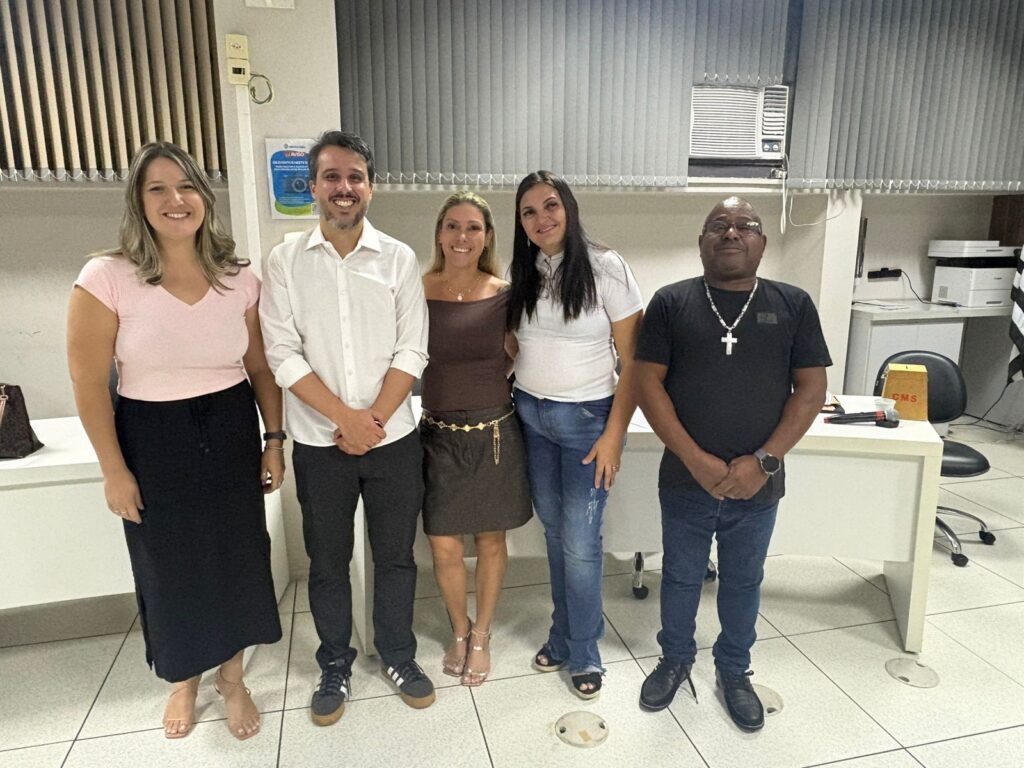 A imagem mostra cinco adultos posando para uma foto em um ambiente interno, possivelmente um escritório ou sala de reuniões. Eles estão alinhados lado a lado, sorrindo para a câmera, transmitindo um clima amigável e descontraído. Da esquerda para a direita, há uma mulher vestindo uma blusa rosa e saia preta longa, um homem com camisa branca e calça escura, uma mulher com blusa e saia marrons, uma mulher com camiseta branca e jeans azul, e um homem com camiseta preta, jeans azul e um colar com crucifixo. Ao fundo, há uma mesa branca com uma caixa amarela sobre ela, onde está escrito "CMS". Também é possível ver uma cadeira de escritório preta, uma impressora e uma janela com persianas verticais. Na parede, há um aviso escrito em português, começando com "AVISO" e "OS EVENTOS". A iluminação é clara, típica de um ambiente interno bem iluminado. O cenário e a postura das pessoas sugerem que pode ser uma reunião informal ou um momento de confraternização no trabalho.