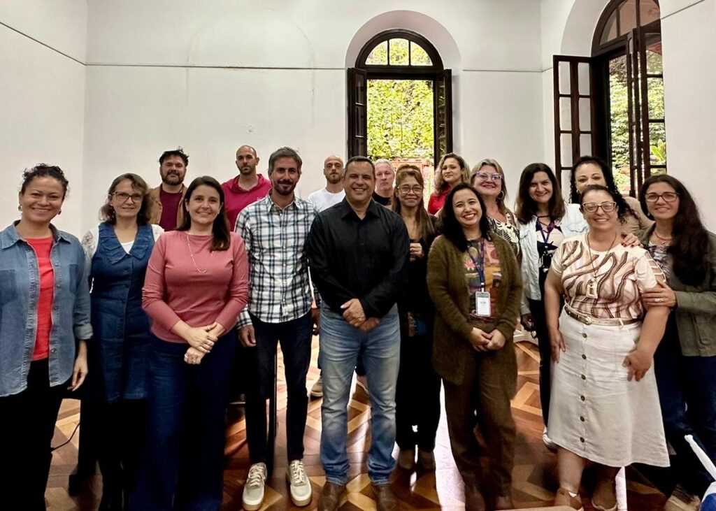 A imagem mostra um grupo de aproximadamente 16 pessoas reunidas em um ambiente interno espaçoso, com paredes brancas e teto alto. O grupo está posicionado em dois níveis, com algumas pessoas agachadas ou sentadas na frente e outras em pé atrás delas. A maioria está sorrindo, transmitindo uma atmosfera amigável e descontraída. As roupas variam entre casuais e semi-formais, incluindo suéteres, camisas, jaquetas jeans e blusas. O chão é de madeira clara com um padrão geométrico, e ao fundo há uma grande porta arqueada de madeira escura que se abre para um espaço externo com vegetação verde. À direita, outra porta aberta revela parte de uma sala adjacente. A iluminação é natural, vinda das portas abertas, complementada pela luz interna. O cenário sugere um encontro de equipe ou grupo de trabalho em um prédio antigo ou institucional.