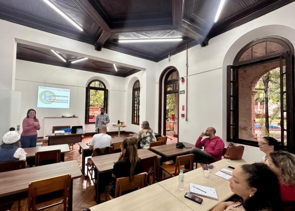 A imagem mostra uma sala ampla com arquitetura clássica, incluindo paredes brancas, teto com painéis de madeira escura e janelas altas e arqueadas que permitem a entrada de luz natural e oferecem vista para um jardim verdejante. No interior da sala, há uma apresentação em andamento. À esquerda, uma mulher vestindo uma camiseta rosa está em pé, gesticulando enquanto fala para o grupo. À direita, um homem de camisa xadrez está próximo a uma tela onde é projetado um slide com o título "TURISMO RURAL" e o subtítulo "OPORTUNIDADES DE EMPREENDIMENTOS", além das palavras "PIRACICABA" e "SAÍDA". Na sala, cerca de oito a dez pessoas estão sentadas em mesas de madeira, de frente para os apresentadores, prestando atenção à palestra. Sobre as mesas, há cadernos, canetas, garrafas de água e um celular. A iluminação é uma combinação de luz natural que entra pelas janelas e luz artificial do teto, criando um ambiente claro e convidativo. O cenário sugere que se trata de um workshop ou curso sobre turismo rural, provavelmente em um contexto educacional ou comunitário, com foco em oportunidades de negócios ou empreendedorismo na área do turismo rural.
