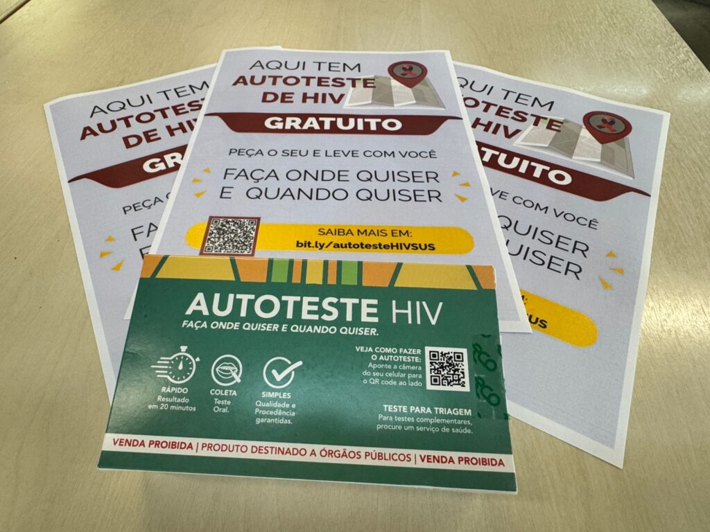 A imagem mostra materiais informativos sobre autotestes de HIV gratuitos, provavelmente distribuídos pelo sistema público de saúde no Brasil. Em destaque, há uma caixinha verde e amarela com a inscrição "AUTOTESTE HIV". A embalagem destaca que o teste é rápido (resultado em 20 minutos), feito por coleta oral, simples e com qualidade garantida. Também há um QR code para obter mais informações. Atrás da caixa, estão três folhetos idênticos dispostos em leque. No topo dos folhetos, há um texto em letras grandes e vermelhas que diz "AQUI TEM AUTOTESTE DE HIV GRATUITO". Abaixo, há um mapa ilustrado com um marcador em forma de fita vermelha, e um QR code para acesso rápido a mais informações. O folheto incentiva as pessoas a pegarem o autoteste e realizarem o exame onde e quando quiserem, com um link para o site bit.ly/autotesteHIVSUS. O fundo é uma superfície clara, possivelmente uma mesa, e a imagem está bem iluminada, focando nos detalhes dos materiais. A composição transmite a ideia de facilidade e acessibilidade do autoteste de HIV para a população.