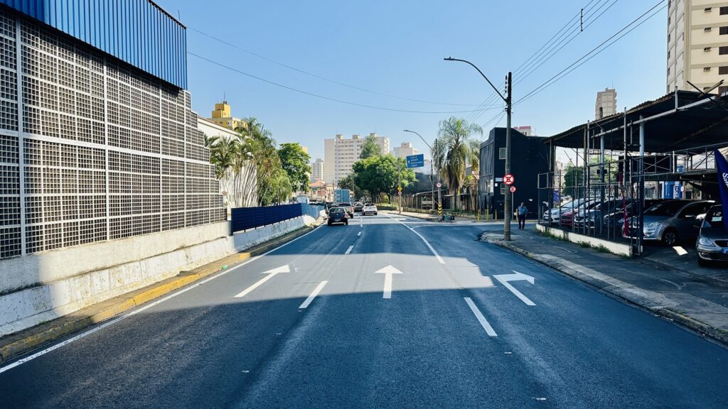 A imagem mostra uma rua larga de uma cidade, provavelmente no Brasil, com três faixas de trânsito bem demarcadas. As setas pintadas no asfalto indicam as direções permitidas para cada faixa: a faixa da esquerda permite seguir em frente ou virar à esquerda, a faixa do meio permite seguir em frente, e a faixa da direita é para virar à direita. À esquerda da imagem, há um grande prédio com um telhado azul e uma fachada feita de blocos de concreto, que parece ser uma construção industrial ou comercial. À direita, há um estacionamento coberto por uma estrutura metálica, com vários carros estacionados. Também há uma pessoa caminhando na calçada próxima a um poste de luz. Ao fundo, é possível ver prédios altos residenciais e muitas árvores, indicando que a área é urbana, mas com bastante vegetação. O céu está claro e azul, sugerindo que a foto foi tirada em um dia ensolarado. No asfalto, estão escritos nomes como "ESALQ", "UNIMEP", "Odontologia" e "FUMEP", que provavelmente indicam direções para instituições de ensino ou unidades específicas próximas. A imagem transmite uma sensação de um dia típico na cidade, com trânsito leve e ambiente organizado.
