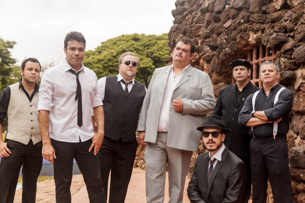 A imagem mostra um grupo de sete homens posando para uma foto profissional ao ar livre. Eles estão vestidos de forma formal e semi-formal, com roupas que vão desde ternos completos até coletes e suspensórios. A paleta de cores é neutra, predominando o preto, branco e cinza, contrastando com o tom terroso da parede de pedra ao fundo e o verde das árvores. Os homens estão organizados em diferentes posições: alguns estão em pé, outros sentados ou agachados. Um homem no centro se destaca por usar um terno cinza claro listrado e um colar com uma cruz, olhando levemente para cima. Outro homem usa óculos escuros e um colete preto, transmitindo uma aparência séria. A iluminação é suave, sugerindo um dia nublado, o que cria sombras delicadas ao redor. O cenário ao fundo é rústico, com uma parede de pedra e vegetação, dando um ar sóbrio e estilizado à foto, que lembra a estética de um grupo musical ou elenco de uma série policial.