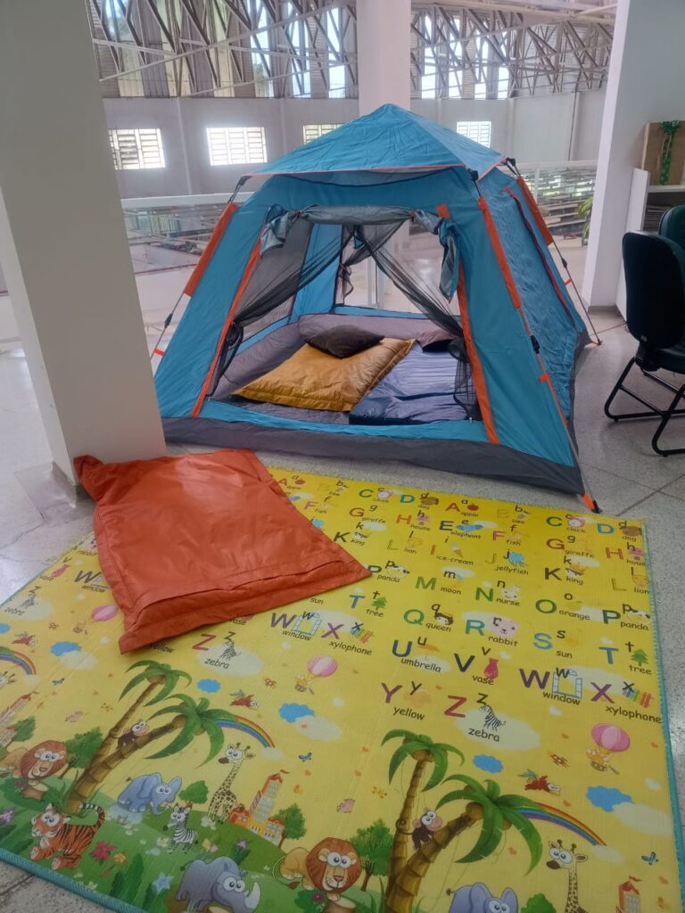 A imagem mostra um ambiente interno amplo e iluminado, onde foi montada uma cena de acampamento. No centro, há uma barraca azul e laranja instalada sobre o piso de azulejos, com dois colchonetes coloridos em seu interior — um amarelo e outro azul. À frente da barraca, um saco de dormir laranja está estendido sobre um tapete infantil ilustrado com o alfabeto, cada letra acompanhada de desenhos e palavras como “A - apple”, “B - ball”, “C - clock”, entre outros. O tapete também traz figuras de animais como leões, girafas e elefantes, além de elementos lúdicos como arco-íris e árvores tropicais.