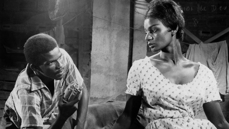 A imagem é uma cena em preto e branco, provavelmente de um filme, que mostra dois jovens negros em um ambiente simples e rústico. O jovem à esquerda está inclinado para frente, olhando fixamente para a jovem à direita, segurando alguns objetos cilíndricos na mão, que parecem ser dinamites ou algo semelhante. Ele veste uma camisa xadrez de mangas curtas. A jovem à direita está com uma expressão séria e um pouco triste, olhando para baixo, vestindo um vestido claro com estampa de pequenos cisnes. O fundo é uma parede de tijolos e concreto, com uma roupa pendurada em um varal e a frase "Amor de Deus" escrita na parede. A iluminação destaca os rostos e as expressões dos personagens, criando uma atmosfera tensa e dramática. A cena transmite uma sensação de confronto ou diálogo intenso em meio a um ambiente humilde.