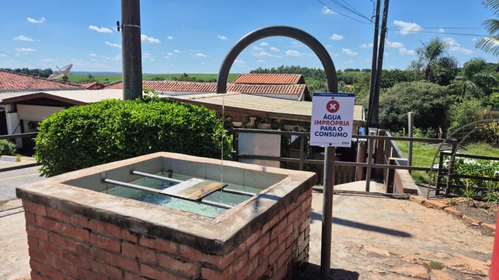 A imagem mostra uma cena ao ar livre em uma área rural ou semi-rural, com um bebedouro ou fonte de água feito de tijolos vermelhos e uma borda de concreto. Uma tubulação metálica curva está despejando um fio de água dentro do bebedouro. A água no reservatório parece limpa, mas há uma placa branca com borda azul e um grande "X" vermelho fixada em um poste próximo, indicando que a água não é própria para consumo, com a frase em português: "ÁGUA IMPRÓPRIA PARA O CONSUMO". Ao fundo, vê-se um céu azul claro com algumas nuvens brancas, casas com telhados de cerâmica laranja, árvores verdes e colinas ao longe. A iluminação forte sugere que a foto foi tirada durante o dia, provavelmente em horário próximo ao meio-dia. O ambiente transmite uma sensação de tranquilidade, mas a placa alerta para o cuidado com a água disponível no local.