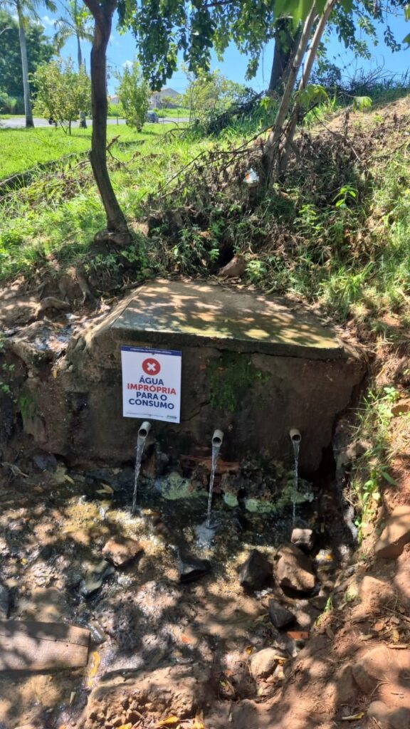 A imagem mostra uma estrutura de concreto embutida em uma encosta, com três canos metálicos de onde sai água. Na frente da estrutura, está fixada uma placa branca com letras pretas que diz: "ÁGUA IMPRÓPRIA PARA O CONSUMO", indicando que a água que sai desses canos não é adequada para beber. Ao redor da estrutura, há terra, pedras e vegetação, sugerindo um ambiente natural, possivelmente uma trilha ou área rural. A iluminação é natural, com sombras de árvores projetadas sobre a estrutura e o solo. Não há pessoas na imagem. A placa também contém pequenos logos no topo e na base, sugerindo que a mensagem foi colocada por uma entidade oficial, provavelmente ligada à saúde pública ou ao município local.