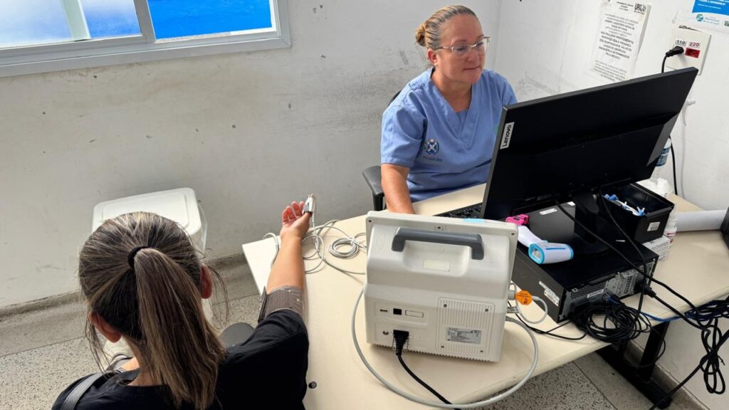 A imagem mostra um ambiente clínico onde uma profissional de saúde está atendendo uma paciente. A profissional, vestindo uniforme azul, está sentada atrás de uma mesa com um computador Lenovo à sua frente. A paciente, vista de costas, está sentada do outro lado da mesa, com um aparelho de pressão arterial no braço e um oxímetro de pulso no dedo. Sobre a mesa há equipamentos médicos, como um monitor e um termômetro digital. Ao fundo, há uma janela que deixa entrar luz natural e algumas cartazes na parede, incluindo um que indica "UPA 24h". A cena transmite um ambiente de atendimento médico organizado e profissional.