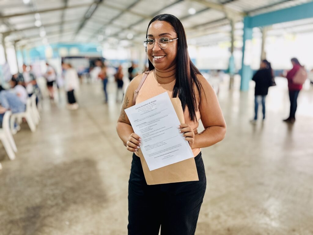 A imagem mostra uma mulher jovem sorridente, segurando um currículo e um envelope marrom, em um ambiente amplo e iluminado, possivelmente um salão de eventos ou uma feira de empregos. Ela usa óculos de armação fina, uma regata cor de pêssego e calças pretas. Seu cabelo é longo e liso, e ela tem uma tatuagem visível no braço direito. Ao fundo, há outras pessoas desfocadas, sentadas ou andando, o que sugere um local movimentado. O currículo que ela segura pertence a Camila Gonçalves de Araújo, uma brasileira solteira nascida em 27/05/1993, residente em Itaim Paulista, São Paulo. Ela tem o ensino médio completo e experiência profissional em vendas externas, estoque e atendimento ao cliente, com habilidades em organização, comprometimento e disposição para aprender. No texto do currículo, Camila solicita uma oportunidade de trabalho, destacando seu compromisso, dedicação e responsabilidade, e se coloca à disposição para uma entrevista imediata.