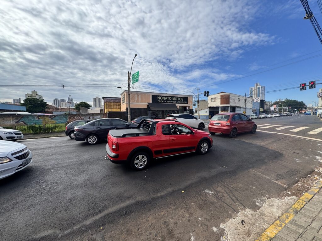 A imagem mostra uma cena urbana em um cruzamento movimentado durante o dia. Em destaque, há uma caminhonete vermelha Volkswagen Saveiro trafegando pela via. Ao redor, há vários veículos, incluindo um Chevrolet Onix branco, um sedã cinza escuro, um SUV branco e um Chevrolet Corsa vermelho. O céu está parcialmente nublado, com nuvens brancas e cinzas contrastando com o azul. Na cena, é possível ver placas de trânsito verdes indicando direções para "RETORNO", "B. Alto", "Sorocaba" e "Tietê", sugerindo que a localização é no estado de São Paulo, Brasil. Também há uma placa de limite de velocidade indicando 50 km/h. No fundo, há estabelecimentos comerciais, incluindo uma loja chamada "NONATO MOTOS PEÇAS E SERVIÇOS" com um número de telefone visível (98133-5363) e outra placa com a palavra "BATERIAS". A foto foi tirada em um ângulo amplo, mostrando a rua asfaltada e as construções ao redor, com uma perspectiva que destaca o movimento do carro vermelho em primeiro plano. A cena transmite o cotidiano típico de uma cidade brasileira, com veículos em trânsito e comércio local ao fundo.