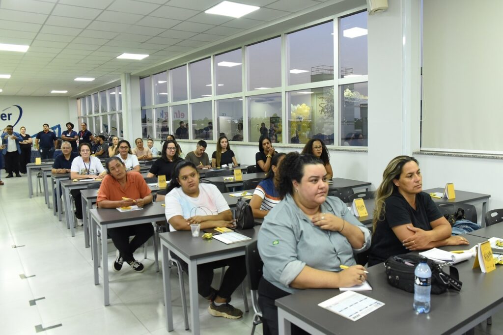 A imagem mostra uma sala de aula ou um ambiente de treinamento corporativo, com várias pessoas sentadas em carteiras organizadas em fileiras. A maioria dos presentes são mulheres, vestidas de forma casual, concentradas em uma apresentação que está acontecendo fora do campo da câmera, provavelmente à direita. Sobre as mesas há cadernos, canetas, garrafas de água e pequenos triângulos amarelos, que podem ser sinalizações ou marcadores. O ambiente é moderno, com teto rebaixado em formato de grade e iluminação de LED embutida. À direita, há uma grande parede de janelas que permite a entrada de luz natural e mostra, do lado de fora, balões amarelos e brancos, indicando que pode haver alguma comemoração ou evento especial acontecendo. Ao fundo, à esquerda, um grupo de pessoas uniformizadas em camisas azuis escuras está reunido próximo a uma parede com uma parte de logo visível, contendo as letras "er". A atmosfera geral é de concentração e aprendizado, em um espaço bem iluminado e organizado.