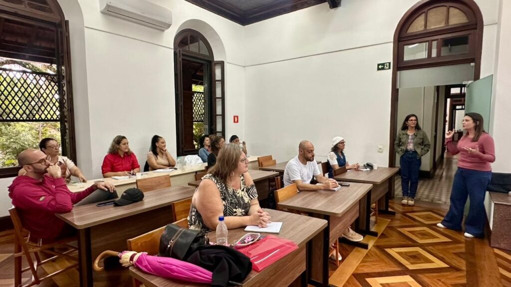 A imagem mostra um grupo de pessoas em uma sala de aula ou reunião, com um ambiente formal e bem iluminado. Há cerca de onze adultos, sendo que a maioria está sentada em carteiras de madeira, enquanto duas mulheres estão em pé, aparentemente conduzindo a atividade ou palestra. A mulher em destaque está usando uma blusa rosa e jeans, gesticulando enquanto fala. A sala possui janelas grandes e arqueadas com molduras escuras, paredes brancas e um piso de madeira com padrão geométrico próximo à porta. Algumas pessoas estão anotando em cadernos, outras apenas escutando atentamente. Sobre as carteiras, há objetos pessoais como cadernos, canetas, garrafas de água e bolsas. O ambiente sugere uma reunião, aula ou workshop, com uma atmosfera de atenção e aprendizado.