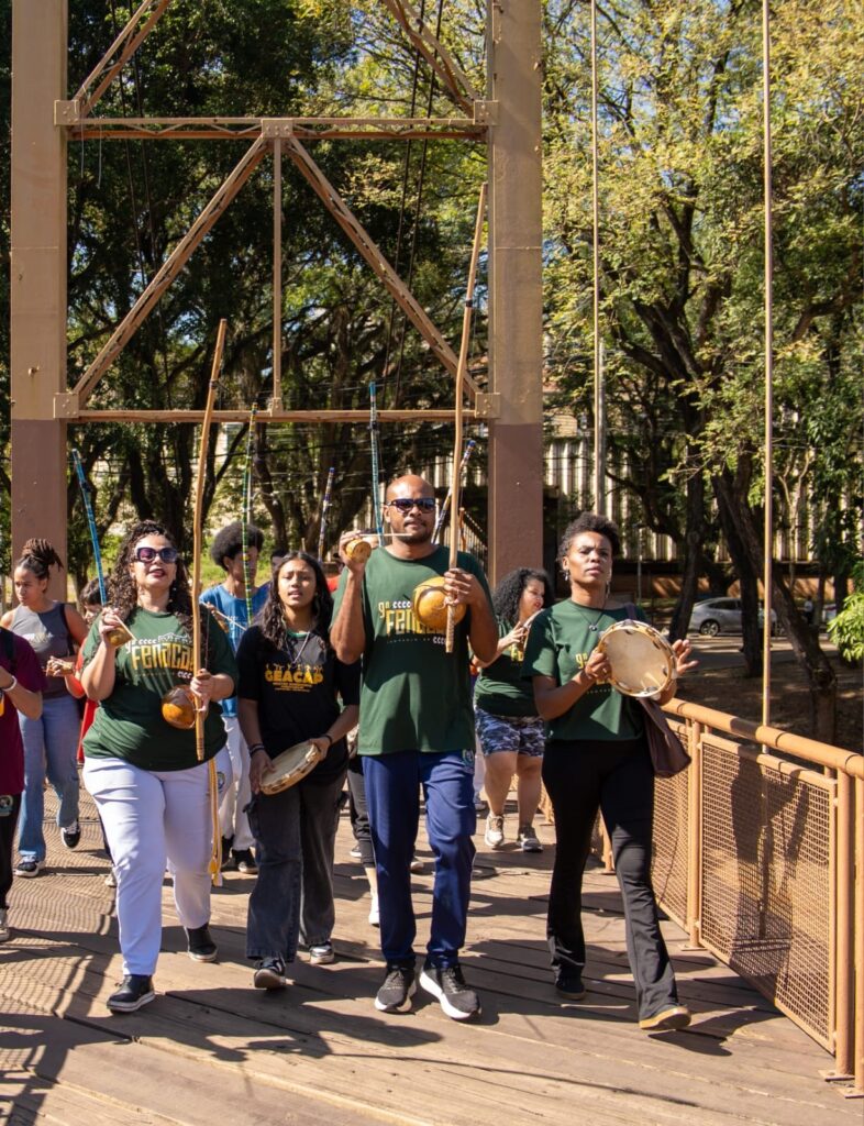 A imagem mostra um grupo de pessoas caminhando por uma ponte suspensa de madeira, enquanto tocam instrumentos tradicionais de capoeira. Na frente, há quatro indivíduos em destaque: duas mulheres e um homem usando camisetas verdes com a inscrição "GEACAP" e "FENACAP", tocando berimbaus e pandeiros, instrumentos típicos da capoeira. Atrás deles, outras pessoas também participam da caminhada, algumas usando camisetas brancas. O cenário é iluminado pela luz do sol, que cria sombras marcantes sobre as tábuas da ponte. Ao fundo, é possível ver árvores verdes e a estrutura metálica da ponte. A imagem transmite uma atmosfera de cultura, tradição e união, provavelmente durante um evento ou celebração relacionada à capoeira.