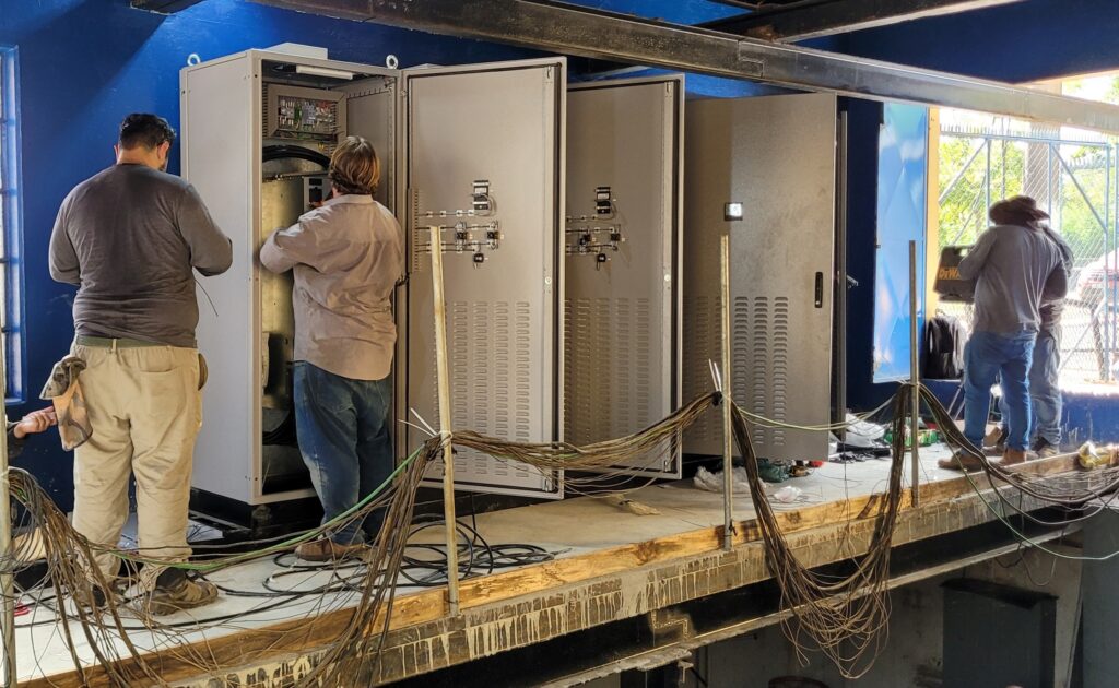 A imagem mostra três técnicos masculinos trabalhando em uma série de painéis elétricos industriais grandes, provavelmente painéis de controle ou de distribuição de energia. Eles estão posicionados sobre uma plataforma de concreto elevada dentro de um ambiente industrial com paredes azuis. Dois dos técnicos estão concentrados na manutenção ou instalação dentro dos painéis abertos, enquanto o terceiro, usando um chapéu de aba larga, caminha para longe dos equipamentos em direção a uma área externa iluminada pela luz do dia. No chão, há vários cabos elétricos grossos e enrolados, indicando um trabalho complexo e em andamento. O ambiente é iluminado tanto por luz artificial quanto pela luz natural que entra pela abertura na parede. A cena transmite a ideia de um trabalho técnico especializado em eletricidade ou automação industrial.