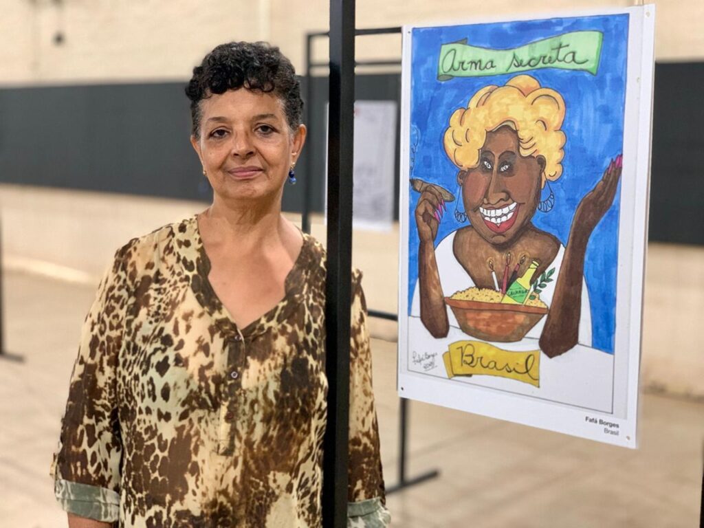 A imagem mostra uma mulher de cabelos curtos, escuros e cacheados, vestindo uma blusa de estampa de oncinha e brincos pequenos azuis. Ela está ao lado de um poste preto, olhando diretamente para a câmera com um leve sorriso. Ao lado dela, há uma ilustração colorida em um painel branco. A ilustração apresenta uma caricatura de uma mulher loira, sorridente, com unhas pintadas de rosa, segurando um charuto. Na frente da caricatura, há uma tigela contendo uma garrafa rotulada como "Cachaça", velas e alimentos. Acima da caricatura, há uma faixa verde com o texto "Arma secreta" e abaixo uma faixa amarela com a palavra "Brasil". Na parte inferior, está o nome "Fafá Borges" e novamente a palavra "Brasil". O ambiente parece ser interno, com uma parede bege ao fundo e uma iluminação suave, sugerindo que a foto foi tirada em uma galeria ou espaço de exposição. A composição da imagem destaca tanto a mulher quanto a ilustração, criando um equilíbrio visual.