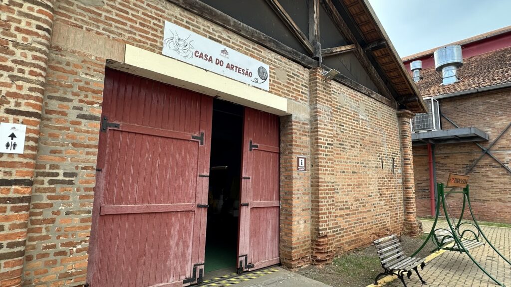 A imagem mostra a entrada de um edifício chamado "Casa do Artesão", que faz parte do Engenho Central, um ponto turístico em Piracicaba. A construção é feita com tijolos aparentes, com uma porta dupla de madeira pintada em tom avermelhado, parcialmente aberta, permitindo ver o interior escuro. Acima da porta, há uma placa branca com o nome "Casa do Artesão" e ilustrações que remetem ao trabalho artesanal, como mãos moldando barro e um novelo de lã. À esquerda da porta, há uma pequena placa indicando banheiro, e à direita, uma placa com a letra "i", que simboliza informações turísticas. Na frente do prédio, há um banco de madeira e uma estrutura metálica verde, possivelmente um balanço ou brinquedo. O chão é parcialmente pavimentado com pedras intertravadas e parcialmente de terra, com vegetação ao redor. Ao fundo, é possível ver outras construções também de tijolo e elementos industriais, como chaminés e dutos. O céu está nublado, sugerindo um dia cinzento. No geral, a imagem transmite um ambiente rústico e histórico, típico de um espaço dedicado à cultura artesanal e turismo.