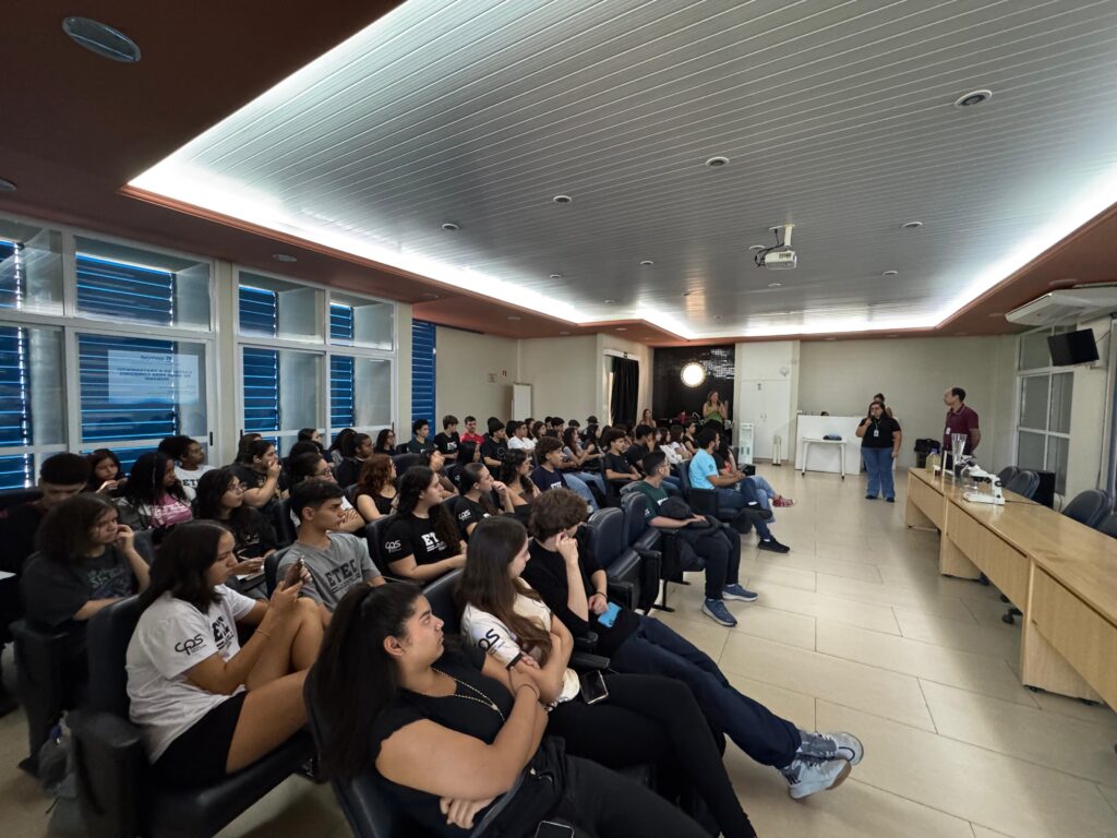 A imagem mostra uma grande sala de aula ou auditório repleto de estudantes sentados em cadeiras alinhadas em fileiras, todos voltados para a frente, onde dois palestrantes estão em pé, aparentemente conduzindo uma apresentação. A maioria dos estudantes usa camisetas com logos da ETEC e CPS, indicando que se trata de uma escola técnica vinculada ao Centro Paula Souza, no Brasil. Na frente da sala, há uma mesa longa com equipamentos de laboratório, incluindo um microscópio e vidrarias, sugerindo que a palestra ou aula está relacionada a ciências, possivelmente química ou biologia. No fundo, há uma janela com persianas azuis e uma placa que menciona "semae" e "Captação e Tratamento de Água para Consumo", o que indica que o tema da apresentação pode estar ligado ao tratamento de água. A iluminação é clara e uniforme, vinda do teto, e o ambiente parece moderno e bem equipado. Os estudantes estão atentos, alguns anotando, demonstrando interesse na palestra. O clima geral é de aprendizado e concentração.
