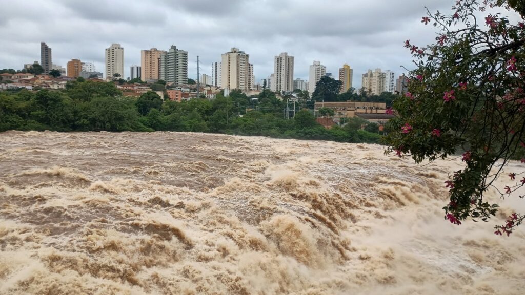 A imagem mostra um rio muito agitado e turvo, com águas marrons e cheias de sedimentos, provavelmente em época de cheia ou enchente. A correnteza está intensa, com muitas ondas e espuma, sugerindo uma força da água muito grande, possivelmente uma cachoeira ou um trecho de corredeira. Ao fundo, vê-se uma cidade com vários prédios residenciais e comerciais, incluindo alguns edifícios altos. A vegetação verde densa separa o rio da área urbana. O céu está nublado, com nuvens cinzentas que indicam tempo fechado ou chuvoso. Em um dos prédios ao fundo, está escrito "BOYES". A imagem transmite uma sensação de força da natureza contrastando com a presença da cidade ao fundo.
