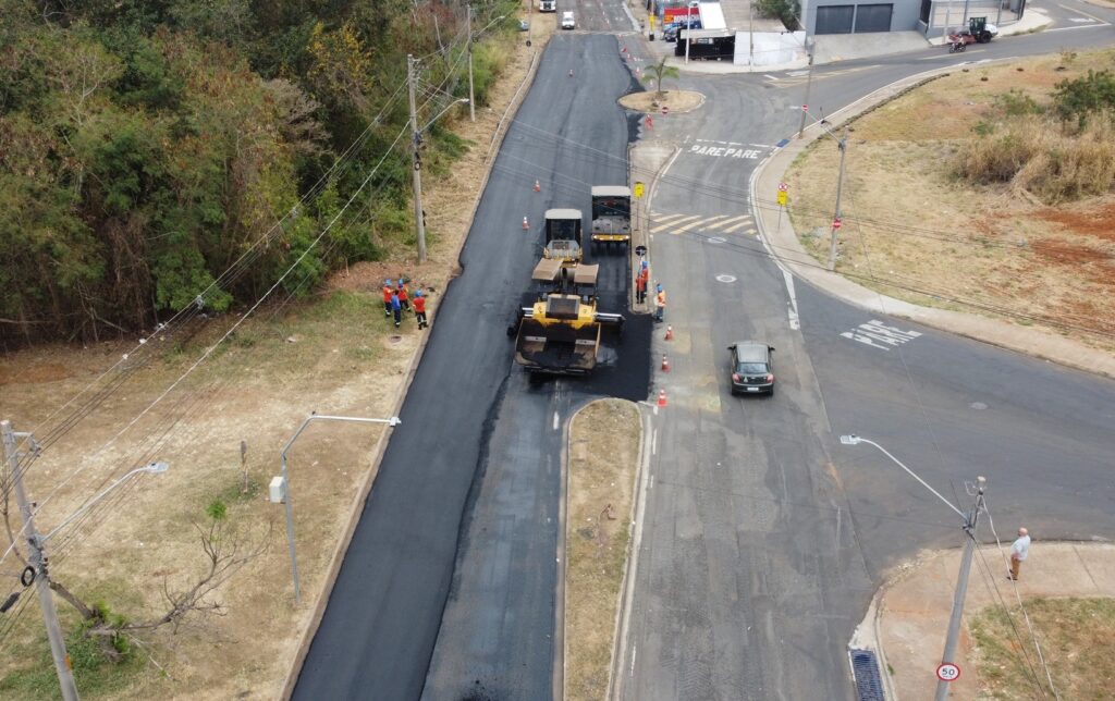 A imagem mostra uma obra de pavimentação asfáltica em um cruzamento de três vias, vista de cima, provavelmente capturada por um drone. O asfalto novo, de cor preta e brilhante, contrasta fortemente com o pavimento antigo, mais claro e desgastado. No centro da imagem, há uma máquina pavimentadora amarela e preta, seguida por um rolo compactador menor e um caminhão basculante. Vários cones de trânsito laranja delimitam a área de trabalho. Há cerca de seis a sete operários vestidos com coletes de segurança nas cores laranja e azul, alguns próximos às máquinas e outros na margem da via, em área com grama verde. Um carro escuro trafega pela pista adjacente, que está liberada para o trânsito. No chão, está pintado o aviso "PARE" em várias partes da via, indicando a necessidade de parada, e uma placa de limite de velocidade de 50 km/h está visível numa das laterais. O ambiente ao redor é uma mistura de áreas verdes com árvores densas de um lado e gramado seco do outro, sugerindo uma região urbana ou periurbana. A cena mostra um trabalho em andamento para renovar a pavimentação da via, com sinalização provisória para garantir a segurança dos trabalhadores e do tráfego.