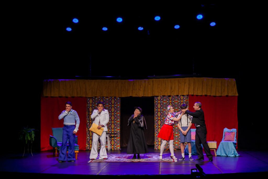 A imagem mostra uma cena teatral com seis atores alinhados no centro do palco, que está decorado com cortinas vermelhas nas laterais, painéis coloridos e um fundo amarelo no topo. O palco é iluminado por luzes azuis, criando um ambiente dramático. Os atores exibem expressões de surpresa ou choque: um homem à esquerda está com a mão na boca, outro segura um envelope amarelo, uma pessoa vestida com traje religioso está com as mãos juntas, uma mulher segura o rosto de um menino, e um homem à direita toca a cabeça do menino. O cenário inclui uma poltrona azul e uma planta à esquerda, e uma cadeira azul com almofada rosa à direita. O chão reflete as luzes coloridas, acrescentando profundidade à cena.