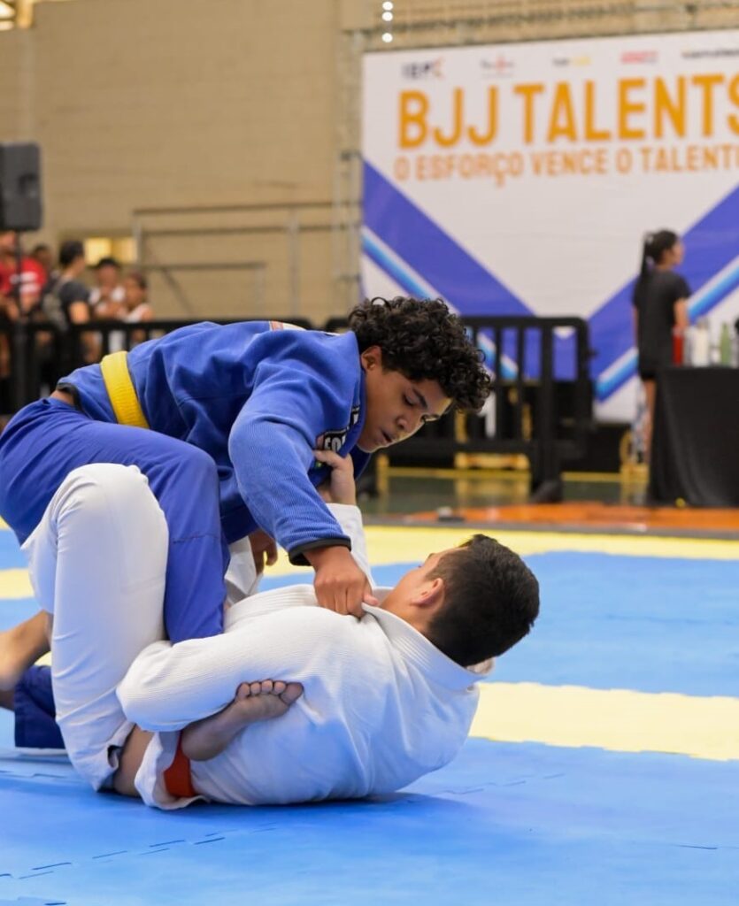 A imagem mostra uma competição de Jiu-Jitsu entre dois jovens atletas. Ambos estão usando quimonos (gis), sendo que um veste quimono azul com faixa amarela e o outro quimono branco com faixa laranja. O atleta de quimono azul está por cima, segurando a lapela do adversário, enquanto o atleta de quimono branco está deitado de costas tentando controlar a perna do oponente. Eles estão sobre um tatame azul com bordas amarelas, típico de competições de artes marciais. Ao fundo, há uma grande faixa laranja com a frase "BJJ TALENTS" e o slogan "O ESFORÇO VENCE O TALENTO", indicando o nome ou o tema do evento. A cena acontece em um ambiente fechado, provavelmente um ginásio ou arena, com algumas pessoas e equipamentos visíveis ao fundo, mas desfocados para destacar os lutadores. A iluminação é clara e uniforme, evidenciando a ação no centro da imagem. A foto foi tirada de um ângulo baixo, destacando a intensidade do combate.