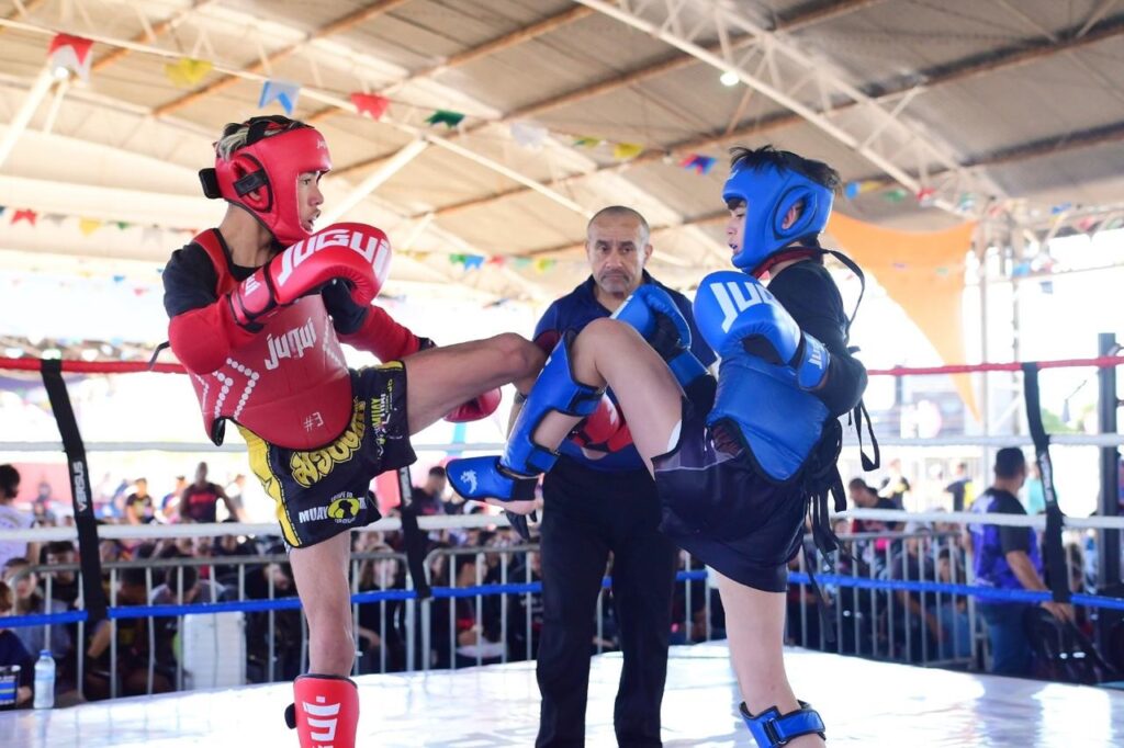 A imagem mostra uma cena vibrante de uma luta de Muay Thai entre dois jovens atletas dentro de um ringue. O lutador à esquerda está usando protetores vermelhos — capacete, luvas e colete — além de shorts pretos com inscrições em amarelo. Ele está no meio de um chute com a perna direita estendida em direção ao adversário. O lutador à direita veste equipamentos azuis — capacete, luvas, colete e caneleiras — e parece estar se preparando para bloquear o golpe. Ambos usam camisas de manga longa pretas por baixo do equipamento de proteção. No centro, um árbitro observa atentamente a luta, vestido com uma camisa polo azul escura e calças pretas. O ringue é cercado por cordas e uma estrutura metálica, e ao fundo é possível ver uma plateia desfocada, indicando um ambiente ao ar livre ou semiaberto, com uma cobertura branca e bandeirinhas coloridas penduradas. A iluminação natural destaca os detalhes dos equipamentos e a intensidade da competição. As marcas "Jugui" aparecem nos equipamentos dos lutadores. A imagem transmite uma sensação de ação, concentração e espírito esportivo.