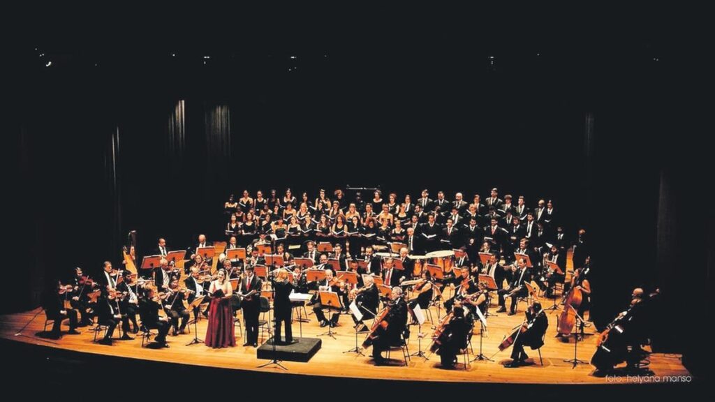 A imagem mostra uma orquestra completa e um coro em uma apresentação no palco de um teatro. A perspectiva é panorâmica e de cima, capturando todo o conjunto. O palco está iluminado com luzes quentes e douradas, que destacam os músicos e cantores contra o fundo escuro da cortina e das paredes do teatro. No centro, está o maestro de costas para a câmera, conduzindo a orquestra que está distribuída em semicírculo. Os músicos tocam instrumentos variados, como violinos, violoncelos, contrabaixos, harpa e percussão. À frente da orquestra, há dois solistas — uma mulher vestida com um longo vestido vermelho e um homem de terno preto — cantando. Atrás da orquestra, há um grande coro organizado em fileiras, com as mulheres vestidas de preto à esquerda e os homens também de preto à direita. A cena transmite uma atmosfera formal e artística, típica de concertos clássicos. No canto inferior direito, há uma pequena legenda com os créditos da foto: "foto: helyana manso".