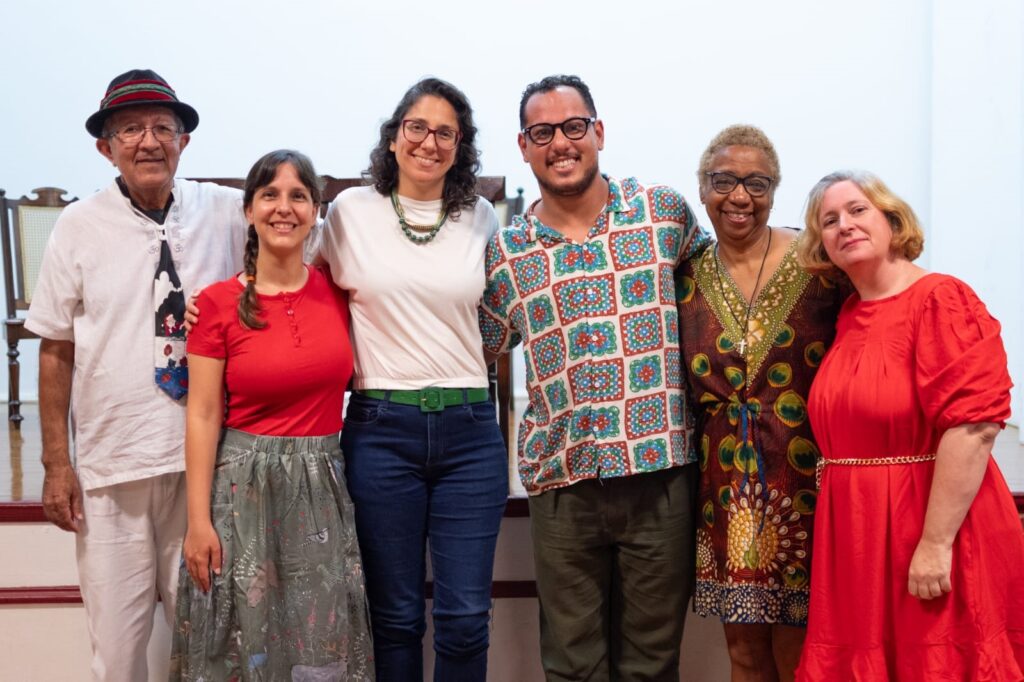 A imagem mostra seis pessoas alinhadas, sorrindo e aparentando estar felizes. Elas são de diferentes idades e etnias, vestindo roupas variadas que refletem estilos pessoais distintos. Da esquerda para a direita, há um homem mais velho usando chapéu preto com faixa vermelha, camisa branca com gravata estampada, e calças claras. Ao lado dele, uma mulher jovem veste uma camiseta vermelha e saia cinza com estampa. A terceira pessoa é uma mulher com óculos, camiseta branca e calça jeans azul, usando um cinto verde e um colar colorido. O quarto é um homem de óculos com uma camisa colorida estampada e calça escura. A quinta pessoa é uma mulher loira de cabelo curto, com vestido escuro estampado com penas de pavão. Por fim, a sexta pessoa é uma mulher loira com vestido vermelho vibrante de mangas bufantes e cinto dourado fino. Eles estão em um ambiente interno, provavelmente em um palco ou plataforma de madeira, e parecem estar em um momento de confraternização ou celebração.