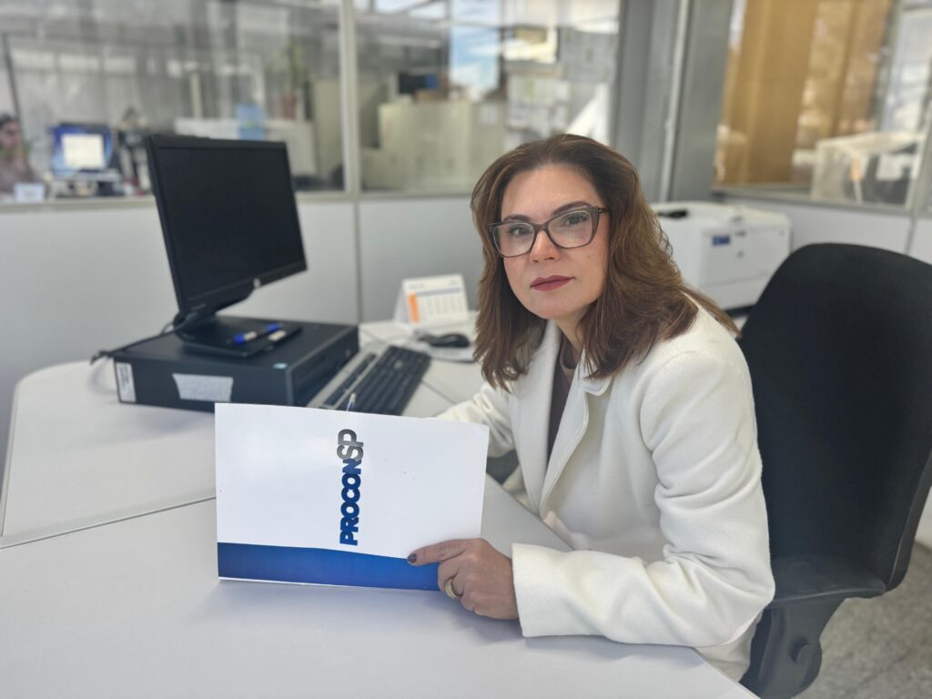 A imagem mostra uma mulher sentada em uma mesa de escritório, segurando uma pasta com a inscrição "PROCONSP". Ela está vestida com um blazer branco sobre uma blusa escura e usa óculos. O ambiente parece ser um escritório moderno, com divisórias de vidro ao fundo, um computador, teclado e uma impressora visíveis na mesa. A iluminação é suave e natural, destacando a mulher em foco, enquanto o fundo está desfocado. A expressão dela é séria e profissional, sugerindo um ambiente de trabalho formal, possivelmente relacionado ao órgão de proteção ao consumidor de São Paulo, o PROCONSP.