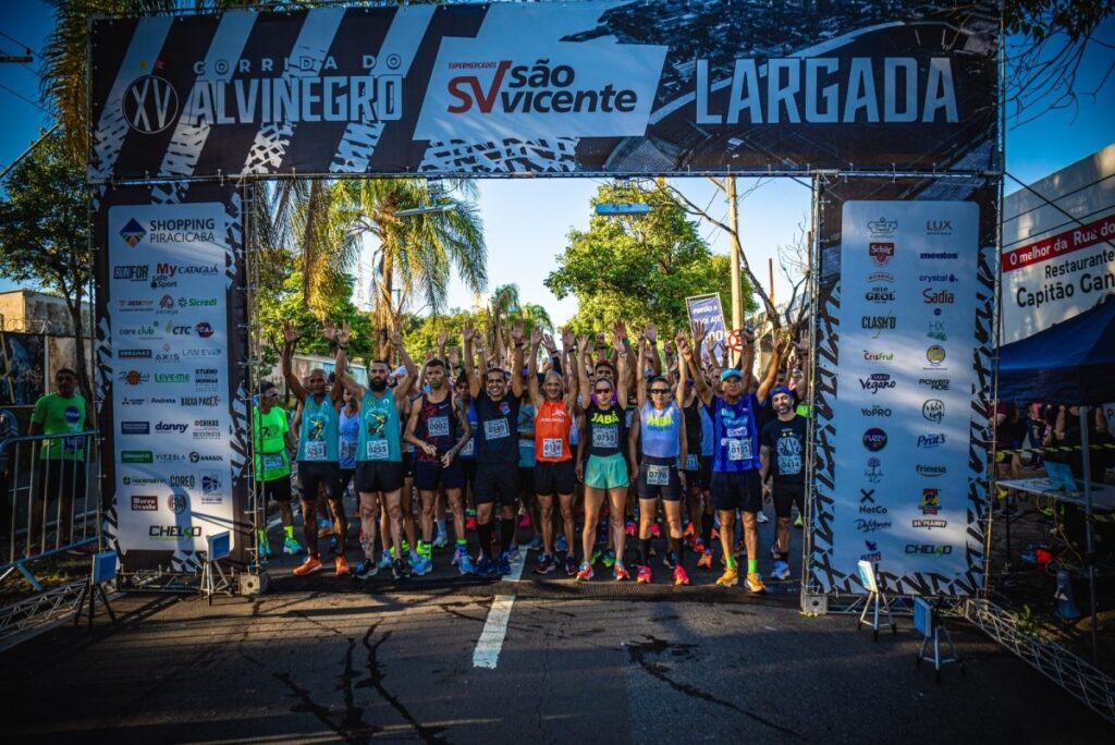 A imagem mostra o momento de largada da XV Corrida do Alvinegro, um evento esportivo de corrida de rua. Vários corredores estão posicionados sob uma estrutura metálica que serve como linha de largada, com os braços levantados, demonstrando entusiasmo e energia. Eles vestem roupas esportivas coloridas e têm números de identificação presos ao peito. A estrutura da largada é bastante decorada com diversas marcas patrocinadoras, como "Supermercados São Vicente", "Shopping Piracicaba", "Sadia", "Mentos", entre outras, evidenciando o apoio comercial ao evento. Acima da estrutura, há uma faixa com o nome da corrida e a palavra "LARGADA" em destaque. O cenário é uma rua asfaltada, iluminada pela luz do sol, sugerindo que a corrida ocorre durante o dia, possivelmente pela manhã ou fim de tarde, devido às sombras longas. Ao fundo, vê-se vegetação com árvores e palmeiras, além de um prédio branco com uma placa indicando um restaurante chamado "Capitão Gan", que aparece parcialmente. No geral, a imagem transmite uma atmosfera de competição saudável, celebração e organização, com um grupo de atletas prontos para iniciar a corrida em um ambiente urbano e ensolarado.