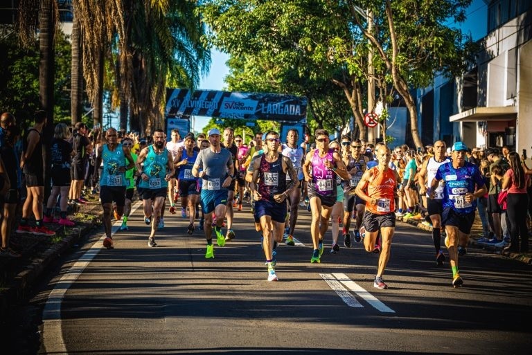 A imagem mostra o momento de largada de uma corrida de rua, com um grande grupo de corredores avançando em direção à câmera. A cena é capturada em um ambiente urbano ou suburbano, com árvores verdes e palmeiras ao longo da rua, sob um céu claro e ensolarado. Muitos corredores vestem roupas esportivas coloridas e números de peito, evidenciando a participação em um evento oficial. No fundo, há uma faixa azul e preta com os dizeres "XV ALVINEGRO", "São Vicente" e "LARGADA", indicando que é a décima quinta edição da corrida Alvinegro, realizada na cidade de São Vicente. A luz do sol cria sombras longas no asfalto, sugerindo que a foto foi tirada pela manhã ou no fim da tarde. A imagem transmite energia, movimento e a emoção do início da competição.