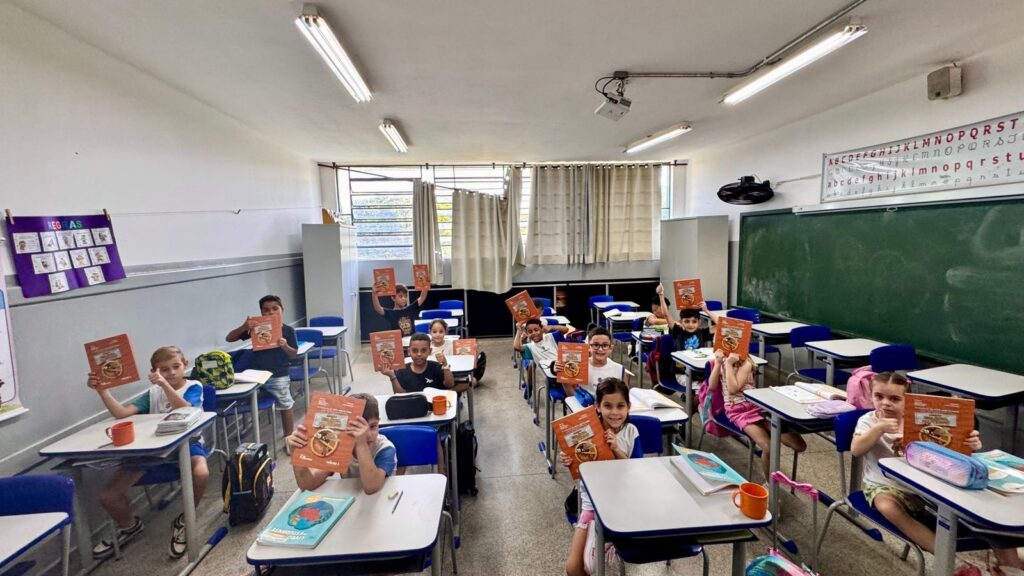 A imagem mostra uma sala de aula iluminada, com cerca de doze crianças em idade escolar sentadas em carteiras azuis e brancas organizadas em filas. As crianças estão olhando para a câmera, muitas segurando livros de capa laranja, que parecem ser livros escolares. Algumas crianças estão sorrindo e fazendo sinal de positivo com o polegar, demonstrando entusiasmo. A sala tem paredes brancas, um quadro verde à direita e uma janela grande com cortinas bege ao fundo, que permite a entrada de luz natural. No teto, há várias luminárias fluorescentes que iluminam bem o ambiente. Na parede à esquerda, há um cartaz roxo com o título "REGRAS" e desenhos que provavelmente ilustram normas de comportamento. Acima do quadro, há uma faixa com o alfabeto em letras maiúsculas e minúsculas. As carteiras estão organizadas de forma a permitir que as crianças fiquem de frente para o professor ou para o quadro, e nelas há mochilas, estojos e copos coloridos. O ambiente transmite uma sensação de aprendizado ativo e descontraído, com as crianças aparentando estar felizes e engajadas.
