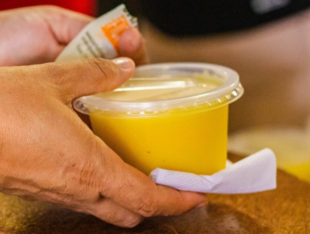 A imagem mostra as mãos de uma pessoa segurando um pequeno recipiente plástico transparente contendo uma substância amarela, que parece ser algum tipo de molho ou sobremesa cremosa. A pessoa está posicionando o recipiente sobre um guardanapo branco. A iluminação é quente e destaca o tom vibrante do conteúdo amarelo. Ao fundo, desfocado, é possível ver uma outra mão segurando um pequeno sachê, possivelmente para adicionar ao conteúdo do recipiente. O foco está nas mãos e no recipiente, com o fundo pouco nítido, sugerindo um ambiente interno, possivelmente um restaurante ou uma lanchonete.