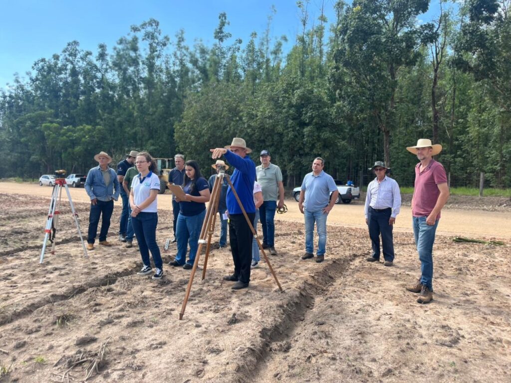 A imagem mostra um grupo de aproximadamente onze pessoas reunidas em um campo aberto de terra batida, possivelmente para uma atividade de topografia ou planejamento agrícola. No centro da cena, um homem com camisa azul de manga comprida e chapéu de palha está apontando para o horizonte, enquanto está ao lado de um equipamento de medição montado em um tripé de madeira, provavelmente um nível de topografia. Ao redor dele, outras pessoas, vestidas com roupas casuais de trabalho, como jeans, camisetas e chapéus, observam atentamente. Uma mulher segura uma prancheta e parece estar anotando informações. O terreno é seco, com marcas que indicam sulcos ou trilhas. Ao fundo, há uma linha densa de árvores verdes sob um céu azul claro e ensolarado. Também é possível ver alguns veículos, incluindo caminhonetes brancas e uma máquina pesada, estacionados ao longo de uma estrada de terra próxima ao campo. A iluminação forte sugere que a foto foi tirada durante o meio do dia. A composição da imagem destaca a interação do grupo em seu ambiente de trabalho ao ar livre, com todos focados na direção apontada pelo homem no centro.