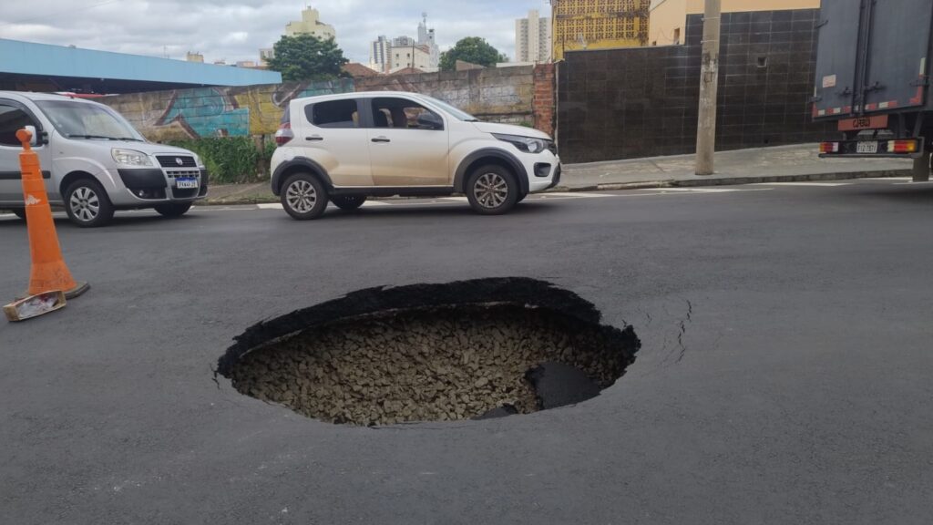 A imagem mostra uma grande cratera ou buraco profundo no meio de uma rua pavimentada em um ambiente urbano. O buraco está preenchido com pedras britadas e está cercado por asfalto escuro. À esquerda do buraco, há um cone de trânsito laranja, provavelmente para alertar os motoristas sobre o perigo. Ao fundo, é possível ver dois veículos da marca Fiat: uma van Fiorino prata e um carro branco modelo Mobi. Também há um caminhão parcialmente visível à direita da imagem. O cenário ao redor inclui um muro com grafite, um prédio com revestimento escuro e edifícios mais distantes, indicando que a foto foi tirada durante o dia em uma cidade. A situação parece ser um problema de infraestrutura, com a necessidade de reparo na via para garantir a segurança dos veículos e pedestres.