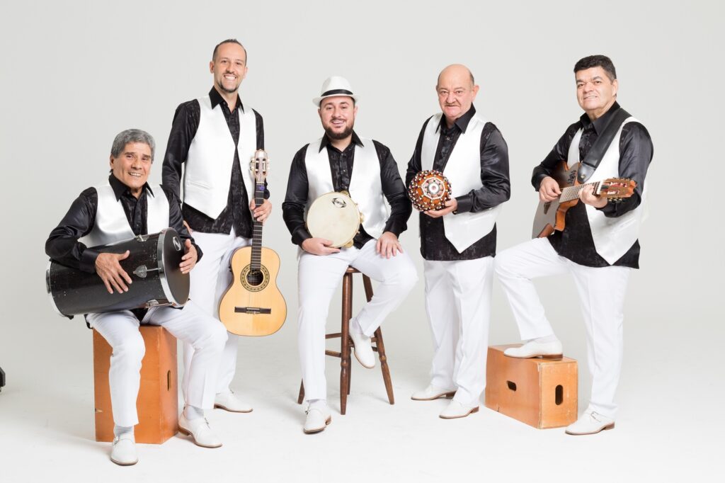 A imagem mostra um grupo musical composto por cinco homens, todos vestidos de forma coordenada com camisas pretas com detalhes em branco, coletes brancos, calças brancas e sapatos brancos. Eles estão posando em um estúdio com fundo branco, transmitindo uma atmosfera profissional e organizada. Da esquerda para a direita: O primeiro homem está sentado em uma caixa de madeira e segura um tambor grande (tan-tan). O segundo homem está em pé, segurando um violão acústico. O terceiro homem está sentado em um banquinho alto, no centro da imagem, usando um chapéu branco estilo fedora e segurando um pandeiro. O quarto homem está em pé, segurando um instrumento de percussão com contas (possivelmente um xequerê). O quinto homem está em pé, com um pé apoiado em uma caixa de madeira, tocando um cavaquinho. Todos estão sorrindo, transmitindo uma energia alegre e descontraída. A imagem destaca a cultura musical brasileira, especialmente gêneros como samba e pagode, evidenciada pelos instrumentos tradicionais. O nome "CONTEMPORANEA" aparece na imagem, possivelmente indicando o nome do grupo.