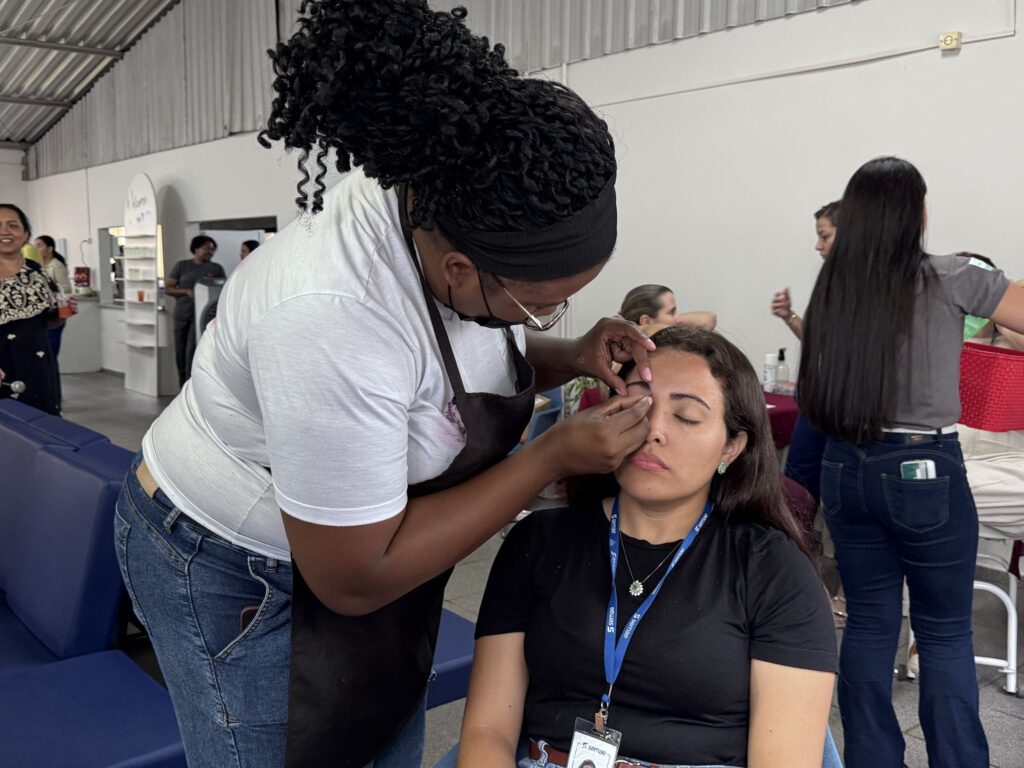 A imagem mostra uma cena em um ambiente interno, possivelmente um salão ou um espaço dedicado a cuidados pessoais. No centro da imagem, uma mulher está realizando um procedimento estético nas sobrancelhas de outra mulher que está sentada com os olhos fechados, aparentando estar relaxada. A profissional usa uma camiseta branca e um avental preto, além de uma faixa preta no cabelo para prendê-lo. A cliente veste uma camiseta preta e um crachá azul com a palavra "semae". Ao fundo, outras pessoas estão presentes, incluindo uma mulher de vestido estampado preto e dourado e outra mulher de camiseta cinza e calça jeans, que parecem estar conversando ou aguardando atendimento. O ambiente é iluminado com luz branca, e há móveis brancos e azuis, sugerindo um espaço organizado e preparado para atender várias pessoas. A atmosfera geral é de um evento ou local voltado para cuidados de beleza e bem-estar.