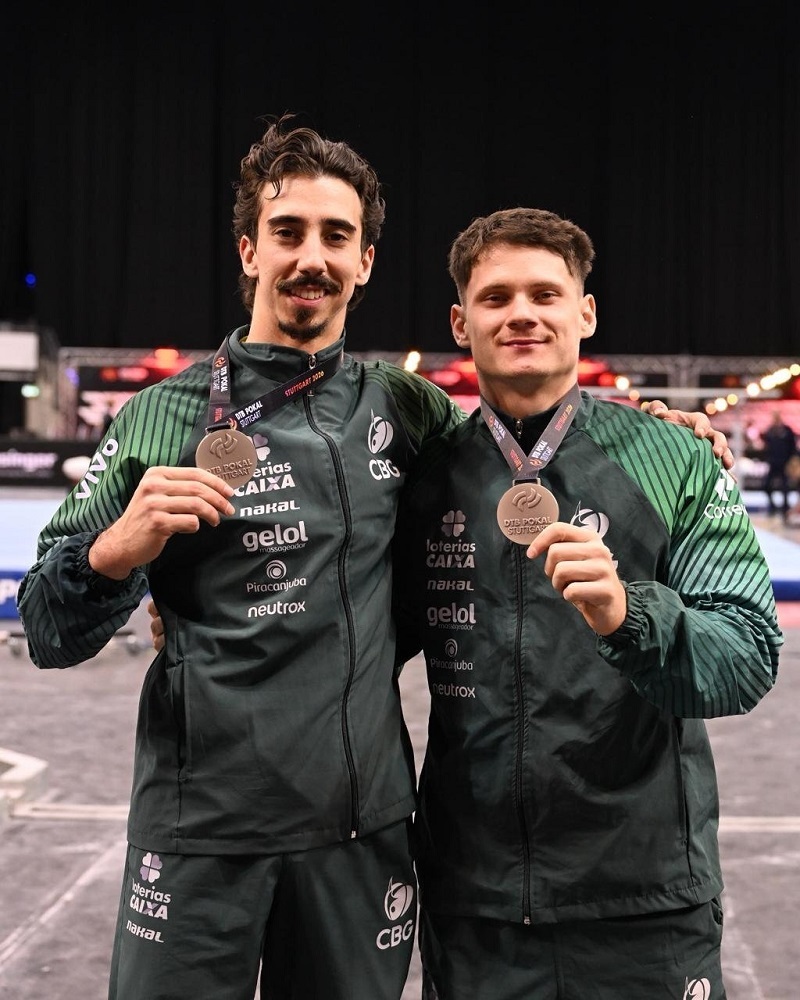 A imagem mostra dois atletas masculinos, provavelmente ginastas brasileiros, posando lado a lado em um ambiente interno, possivelmente uma arena esportiva. Ambos vestem uniformes esportivos verdes escuros com várias logomarcas, incluindo "CBG" (Confederação Brasileira de Ginástica), "Loterias CAIXA", "nakal", "gelol", "Piracanjuba" e "neutrox". Cada um segura uma medalha de bronze pendurada em uma fita preta com a inscrição "DTB POKAL STUTTGART", indicando que participaram de uma competição realizada em Stuttgart, Alemanha. Os atletas estão sorrindo, demonstrando satisfação pela conquista, e estão próximos um do outro, com os braços ao redor das costas, transmitindo camaradagem e espírito de equipe. O fundo está desfocado, destacando os atletas em primeiro plano, e sugere um ambiente esportivo com arquibancadas e iluminação típica de competições indoor. A imagem transmite um momento de celebração e orgulho pela medalha conquistada.