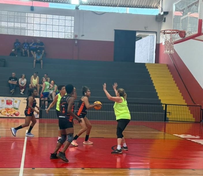 A imagem mostra uma partida de basquete feminino em uma quadra coberta. No centro da cena, uma jogadora vestindo um uniforme preto e laranja segura a bola, enquanto uma adversária, que usa uma camiseta amarela fluorescente, está em posição defensiva com os braços levantados tentando bloquear a jogadora com a bola. Outras duas jogadoras do time de preto e laranja estão próximas, provavelmente se posicionando para receber um passe ou ajudar na jogada. Ao fundo, há arquibancadas com algumas pessoas assistindo ao jogo. O piso da quadra é de madeira clara e há uma área pintada de vermelho próximo à cesta, que está parcialmente visível à direita da imagem. A iluminação é clara e uniforme, típica de ginásios esportivos. A cena transmite a intensidade e movimentação de um jogo em andamento.