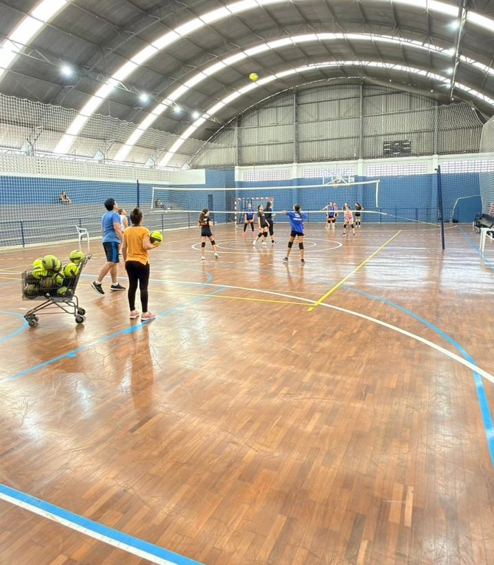 A imagem mostra uma quadra de vôlei indoor com piso de madeira clara e linhas demarcadas em azul, branco e amarelo. No centro da quadra, há uma rede de vôlei estendida. À frente, no canto esquerdo, está um carrinho metálico cheio de bolas amarelas de vôlei. Há também uma bola amarela suspensa no ar, próxima ao teto da quadra. No lado mais próximo da câmera, duas pessoas estão observando a quadra: um homem de camiseta azul e uma mulher de camiseta laranja. Na quadra, várias jogadoras jovens, provavelmente adolescentes, estão distribuídas em posições diferentes, vestindo uniformes esportivos com shorts e camisetas, algumas usando joelheiras. O ambiente é uma quadra esportiva coberta, com um teto alto em formato arqueado feito de metal ondulado, com janelas de vidro ou claraboias que permitem a entrada de luz natural. As paredes ao fundo são azuis, com pilares brancos e uma rede protetora. A cena parece ser um treino ou aula de vôlei, com os observadores possivelmente sendo treinadores ou instrutores.
