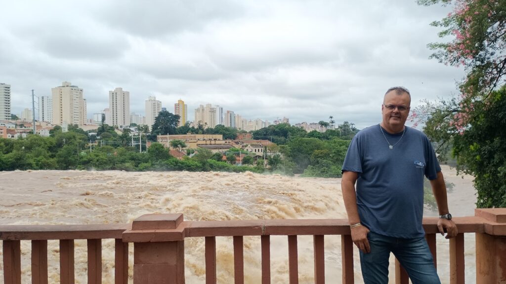A imagem mostra um homem de meia-idade posando em frente a um rio com água turva e agitada, sugerindo uma enchente ou um fluxo intenso de água. Ele está à direita da imagem, vestindo uma camiseta azul, jeans e acessórios como colar, relógio e pulseira. Atrás dele, há um corrimão de cor terracota que separa a área onde ele está do rio. Ao fundo, vê-se uma paisagem urbana com vários prédios altos e muitas árvores verdes, sob um céu nublado e carregado. A palavra "BOYES" aparece em um dos edifícios ao fundo. A cena transmite uma atmosfera de um ambiente urbano próximo a um rio em situação de cheia ou força da água.