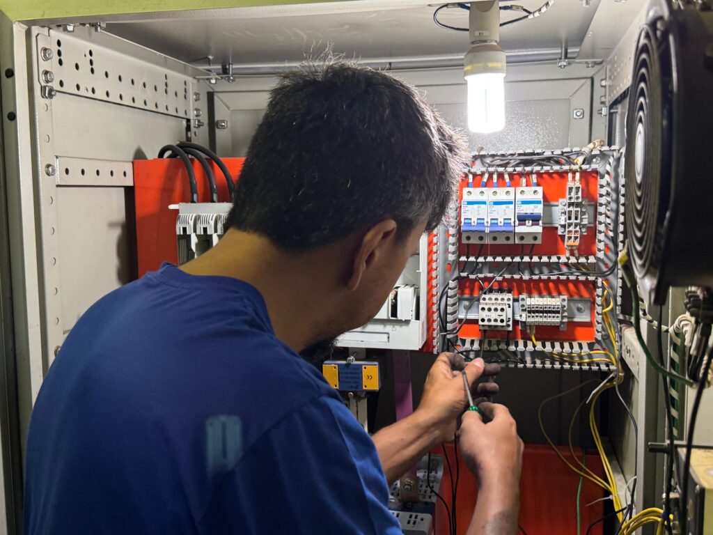 A imagem mostra um técnico trabalhando dentro de um painel elétrico industrial grande. Ele está de perfil, com as costas parcialmente voltadas para a câmera, vestindo uma camiseta azul e segurando uma chave de fenda verde, realizando ajustes ou conexões nos componentes do painel. O interior do painel é iluminado por uma lâmpada localizada na parte superior, que destaca os detalhes dos componentes. No painel, há uma placa laranja com vários dispositivos instalados, como disjuntores identificados como Q1, Q2 e Q3, um contato identificado como KA1, além de diversos terminais e fios pretos e amarelos organizados. Também é visível um grande ventilador preto na lateral do painel. O ambiente sugere um trabalho técnico detalhado e cuidadoso em um sistema elétrico industrial.