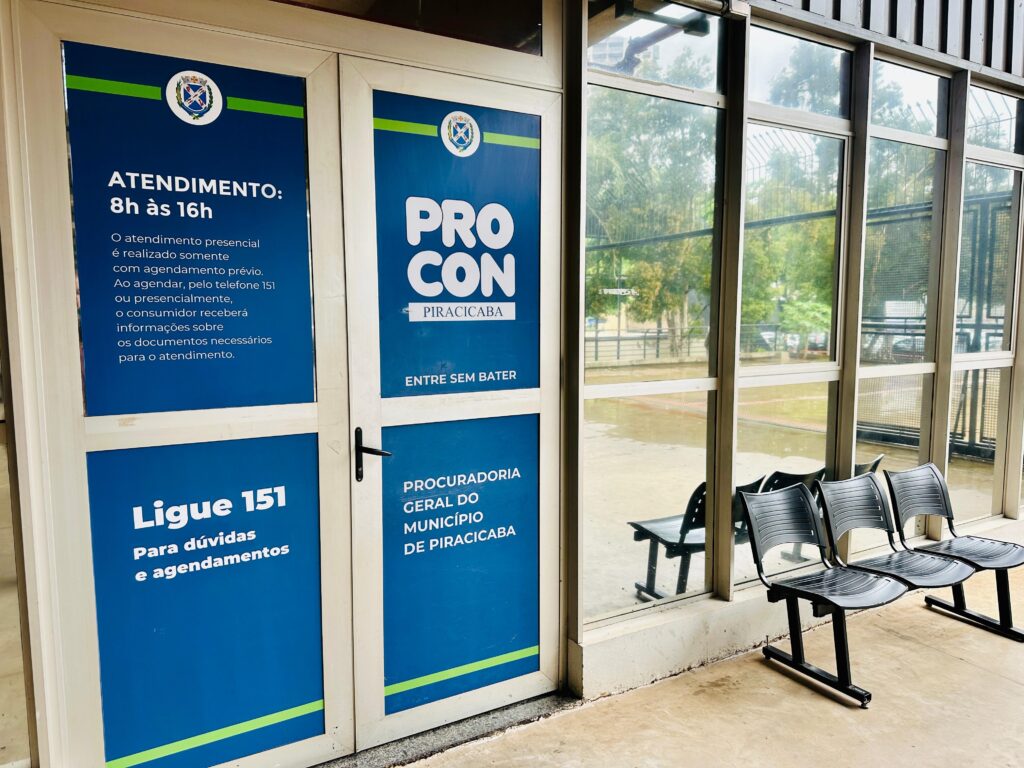 A imagem mostra a entrada do PROCON de Piracicaba, órgão responsável pela defesa do consumidor na cidade. A porta principal é de vidro com moldura branca e apresenta painéis azuis com informações sobre o atendimento. No painel à esquerda, há o horário de funcionamento: das 8h às 16h, e uma orientação importante de que o atendimento presencial só é realizado mediante agendamento prévio, que pode ser feito pelo telefone 151 ou presencialmente. Também informa que, ao agendar, o consumidor receberá detalhes sobre os documentos necessários para o atendimento. Ao lado direito da porta, há uma parede de vidro, atrás da qual estão alinhadas quatro cadeiras pretas, provavelmente para espera dos consumidores. Através do vidro, é possível ver árvores e uma cerca, indicando que o local está em uma área com vegetação externa. No topo da porta, está escrito "PROCON PIRACICABA" e "PROCURADORIA GERAL DO MUNICÍPIO DE PIRACICABA". A frase "ENTRE SEM BATER" está destacada em letras brancas sobre o fundo azul, sugerindo que a entrada é livre, mas controlada pelo agendamento. O ambiente parece organizado e preparado para receber o público, com um layout simples e funcional. A imagem transmite uma sensação de formalidade e seriedade, condizente com o serviço público oferecido.