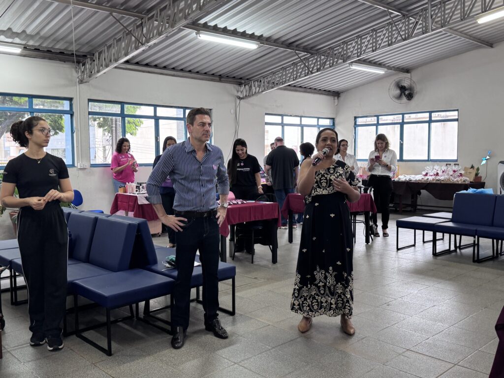 A imagem mostra um evento interno realizado em um amplo salão de estilo industrial, com paredes brancas e piso cinza. O teto é alto, feito de metal ondulado, com luminárias fluorescentes suspensas. Grandes janelas com moldura azul permitem a entrada de luz natural, iluminando o ambiente. No centro da cena, há três pessoas em destaque: uma mulher de vestido longo preto e branco falando ao microfone, um homem ao lado dela com camisa azul listrada e calça jeans, e uma jovem mulher vestindo camiseta preta com a marca "espaçolaser". Ao fundo, há mesas cobertas com toalhas rosa, contendo diversos objetos, possivelmente brindes ou materiais de divulgação. Outra pessoa usa uma camiseta rosa com a marca "Mary Kay". O ambiente sugere um evento corporativo, workshop ou feira, com as pessoas interagindo e prestando atenção na fala da mulher com o microfone. O mobiliário inclui bancos modulares azuis no primeiro plano, que dão um toque moderno ao espaço. A atmosfera é descontraída e profissional ao mesmo tempo.