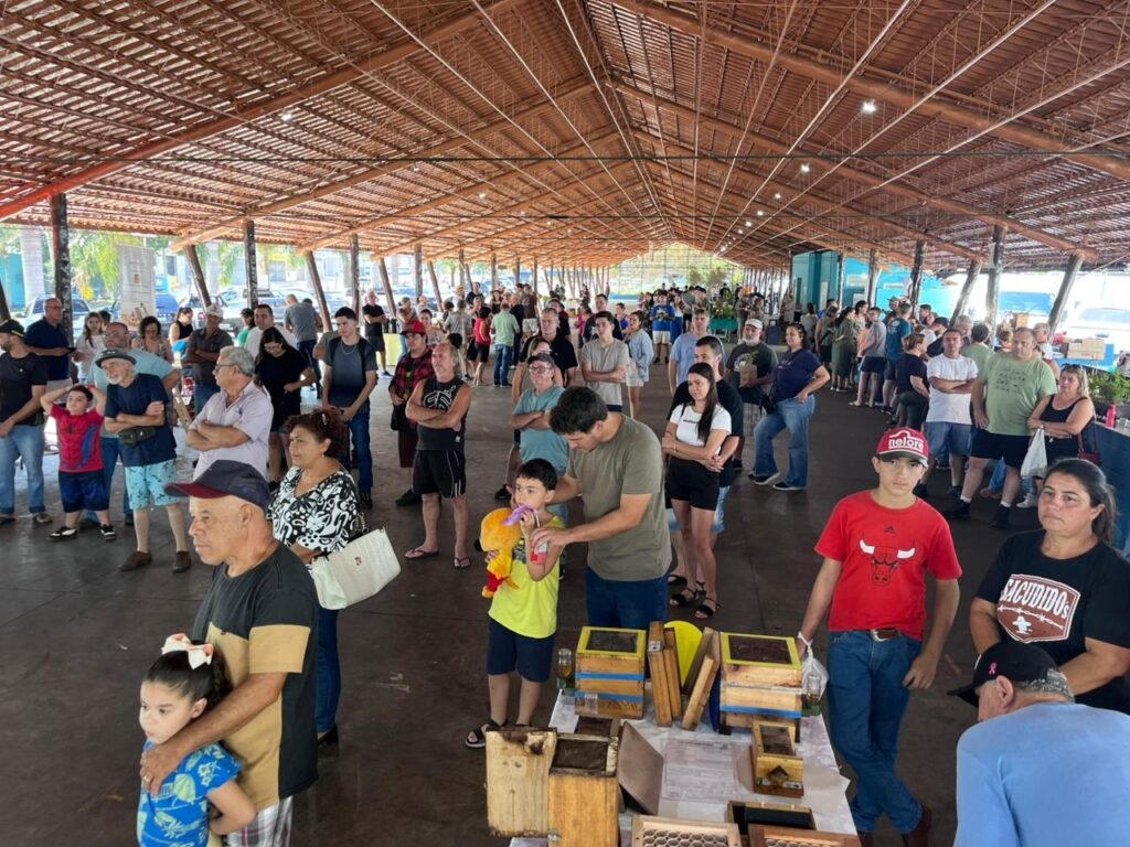 A imagem mostra uma grande quantidade de pessoas reunidas sob uma estrutura de madeira com telhado de palha ou madeira, em um ambiente ao ar livre que lembra uma feira ou evento rural. O local é uma espécie de pavilhão aberto, com muitas pessoas de diferentes idades, algumas observando objetos expostos sobre mesas, que parecem caixas de criação de abelhas (meliponicultura). No primeiro plano, há um homem usando boné vermelho com a palavra "nelore" escrita, e uma camiseta preta com o logotipo do Chicago Bulls. Também é possível ver uma criança segurando um brinquedo amarelo. A multidão está distribuída ao longo do pavilhão, algumas pessoas parecem estar em fila, outras conversando ou olhando as exposições. O ambiente transmite uma atmosfera comunitária e rural, possivelmente relacionada à agricultura ou apicultura, dada a presença das caixas de abelhas e o boné com referência à raça Nelore, comum na pecuária brasileira. A iluminação natural reforça o clima de dia ensolarado.