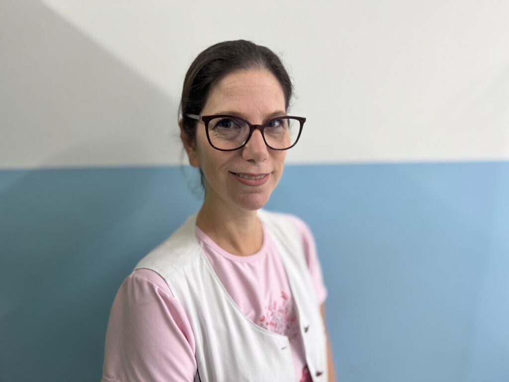 A imagem mostra uma mulher de meia-idade com cabelos castanhos presos para trás, usando óculos de armação escura em estilo gatinho. Ela está vestindo uma camiseta rosa clara por baixo de um colete branco, que lembra um uniforme de profissional de saúde ou educadora. A mulher olha diretamente para a câmera com um sorriso gentil e expressão amigável. O fundo é simples, dividido horizontalmente em duas cores claras: branco na parte superior e azul claro na inferior, com iluminação suave e uniforme. A foto é um retrato médio, com foco nítido no rosto da mulher, transmitindo uma sensação de profissionalismo e simpatia.