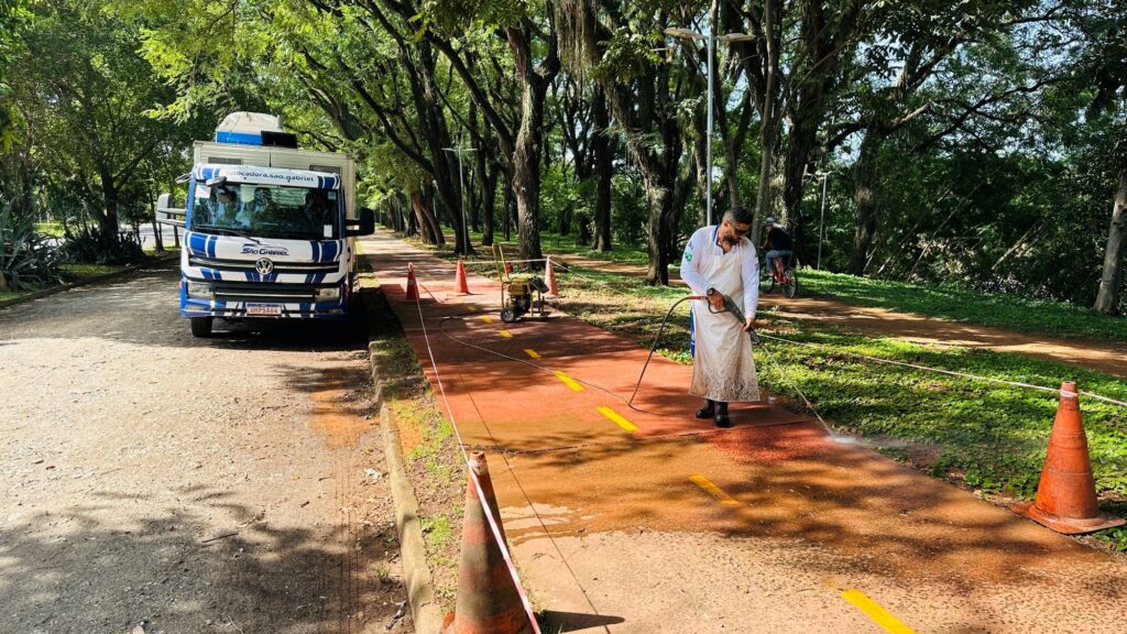 A imagem mostra uma cena ao ar livre em um caminho pavimentado de cor avermelhada, cercado por árvores altas e verdes. No centro da imagem, há um homem usando uma camisa branca de manga longa, calça escura, botas e um avental branco sujo. Ele está utilizando uma lavadora de alta pressão para limpar o caminho, com o jato d'água visível atingindo o chão e deixando a área molhada. À esquerda do homem, está estacionado um caminhão branco da marca Volkswagen, com detalhes em azul e laranja, e inscrições que incluem "@locadora.sao.gabriel" e "SÃO GABRIEL". Na frente do caminhão, há cones de sinalização laranja alinhados ao longo do caminho, delimitando a área de trabalho e conectados por uma corda branca. Ao fundo, uma pessoa está andando de bicicleta, afastando-se da câmera. A iluminação é clara e ensolarada, com sombras das árvores projetadas no chão. A cena transmite uma sensação de manutenção e cuidado do espaço público, em um ambiente natural e arborizado.
