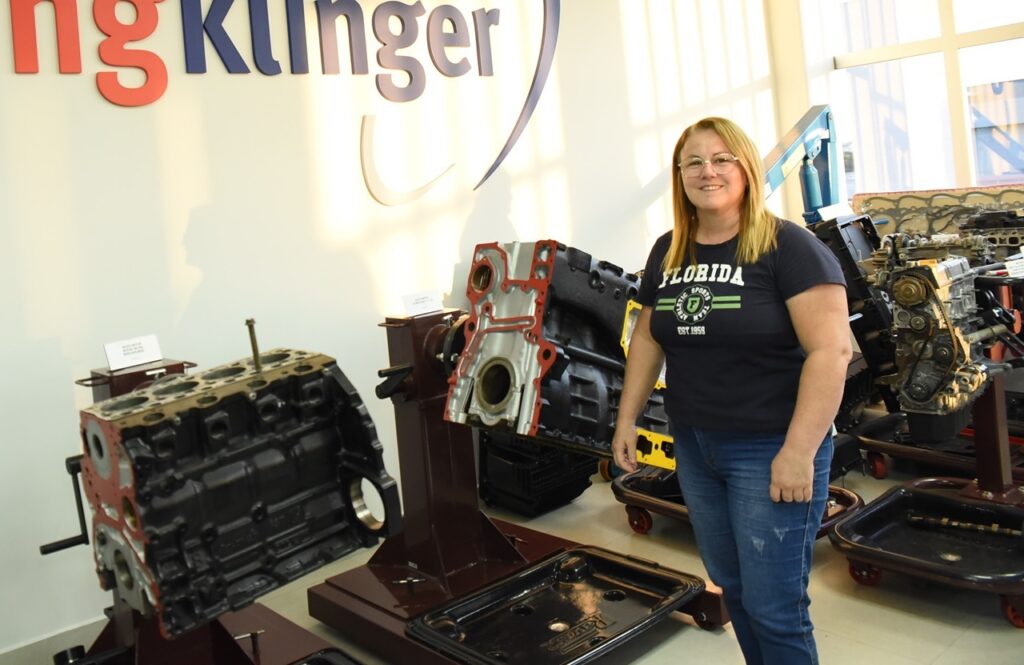 A imagem mostra uma mulher em um ambiente interno que parece ser uma exposição ou showroom de motores. Ela está sorrindo, usando óculos, camiseta azul escura com a palavra "FLORIDA" e jeans azul. Ao redor dela, há grandes blocos de motor expostos em suportes metálicos. Ao fundo, na parede branca, há um logotipo grande com a palavra "Klinger". A iluminação é natural, vinda de janelas grandes atrás dela, o que deixa o ambiente bem claro. A mulher está posicionada ao lado dos motores, o que ajuda a dar uma noção do tamanho das peças expostas. O cenário sugere que se trata de um local relacionado à engenharia automotiva ou uma empresa que trabalha com motores.