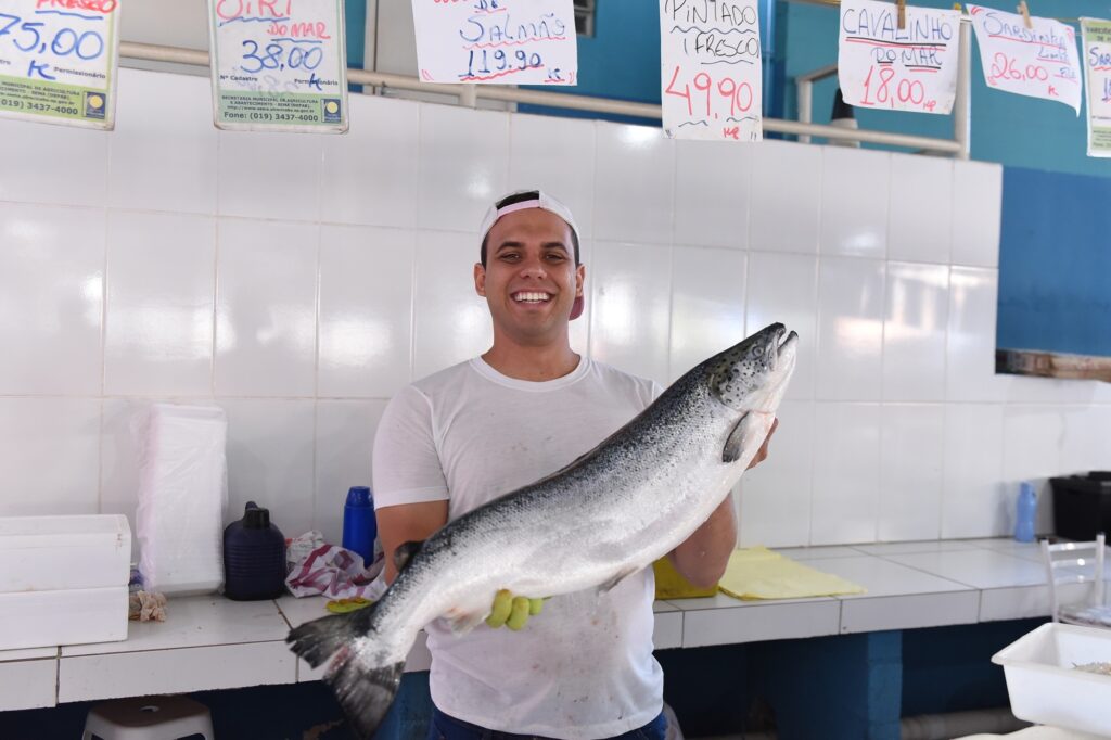 A imagem mostra um homem sorridente, vestindo uma camiseta branca e um boné branco, segurando um grande salmão inteiro na horizontal, à altura do peito. Ele está em um ambiente que parece ser uma peixaria, com paredes revestidas de azulejos brancos e um balcão também revestido com azulejos brancos à sua frente. O homem usa luvas verdes, provavelmente para manusear o peixe com higiene. Acima dele, pendurados, há vários cartazes com preços de peixes escritos à mão, em português, indicando valores por quilo, como "SALMÃO 119,90" e "CAVALINHO DO MAR 18,00". No balcão, há caixas de isopor, garrafas e outros utensílios típicos de um comércio de pescado. O ambiente é bem iluminado, transmitindo uma sensação de limpeza e organização. O homem transmite simpatia e orgulho ao mostrar o salmão, sugerindo que é o vendedor ou proprietário do estabelecimento.