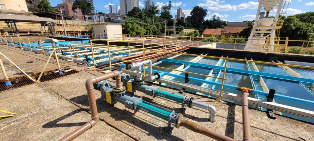 A imagem mostra uma estação de tratamento de água industrial vista de um ângulo alto e amplo. Em primeiro plano, há duas bombas de cavidade progressiva da marca NETZSCH, de cor verde e cinza, instaladas sobre uma base de concreto. Essas bombas estão conectadas a uma rede de tubulações marrons e prateadas. Ao fundo, é possível ver grandes tanques retangulares de decantação ou clarificação, com água escura, divididos por estruturas metálicas azuis e brancas. Em volta dos tanques, há corrimãos amarelos para segurança. Além da área industrial, observa-se uma mistura de elementos urbanos: casas com telhados vermelhos, árvores verdes, prédios altos e uma torre metálica branca. O céu está claro, com poucas nuvens brancas, e a iluminação do sol é intensa, gerando sombras marcantes no chão de concreto. A cena transmite uma atmosfera funcional e técnica, típica de uma instalação de tratamento de água ou esgoto.