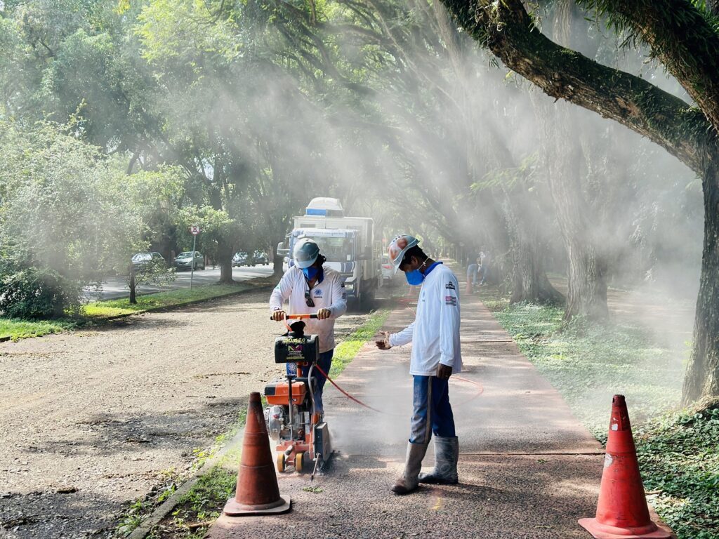 A imagem mostra dois trabalhadores em uma área externa, possivelmente em um parque ou calçada arborizada, realizando um serviço de corte no pavimento. Um dos trabalhadores está operando uma serra elétrica de corte de concreto, que está gerando muita poeira branca no ar, criando uma névoa ao redor. Ambos os trabalhadores estão usando capacetes, máscaras faciais e roupas de proteção, incluindo camisas de manga longa e calças azuis. Há cones de sinalização laranja no chão para delimitar a área de trabalho. Ao fundo, há uma caminhonete branca estacionada e alguns carros passando em uma via próxima. As árvores grandes formam uma espécie de túnel verde, e a luz do sol penetra entre as folhas, contribuindo para o efeito visual da poeira no ar. A imagem transmite uma sensação de trabalho pesado e cuidado com a segurança no local.